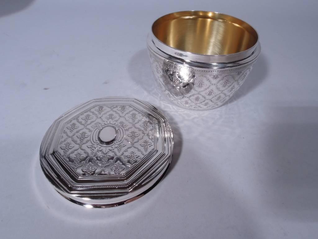 Sterling silver vanity jar. Made by Tiffany & Co. in New York. Bowl has curved sides and flat cover. Acid-etched pattern with two-leaf plant set in scrolled frame. Cover has same set in molded octagonal frame with dentil border. Box and cover