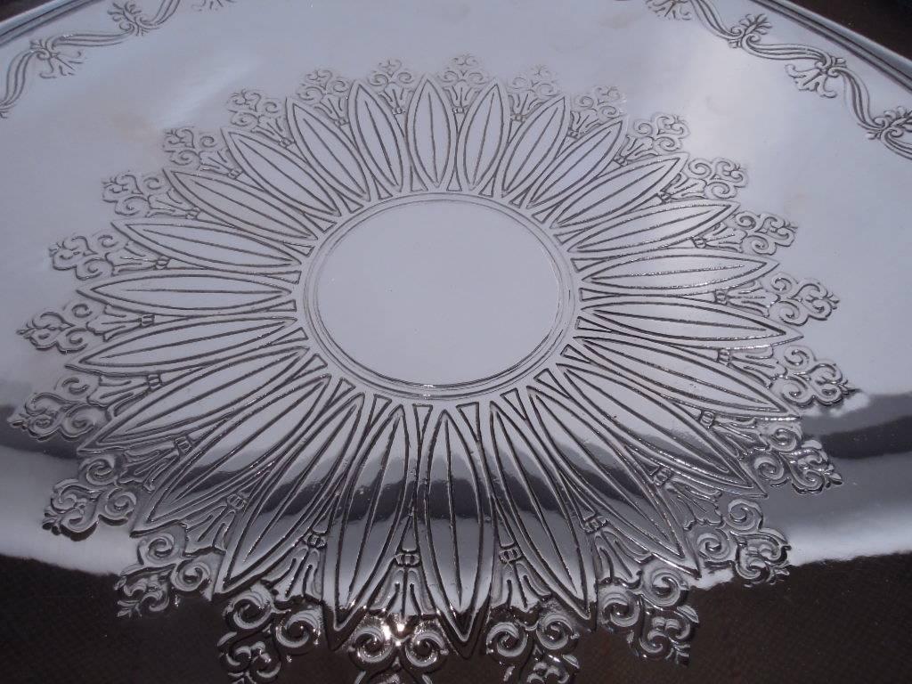 Sterling silver cake plate. Made by Tiffany & Co. in New York, circa 1917. Acid-etched and stylized ornament: Circular frame (vacant) bordered by petals inset with flowers. Scrolled border has alternating palmettes and fleurs-de-lis. Short foot.