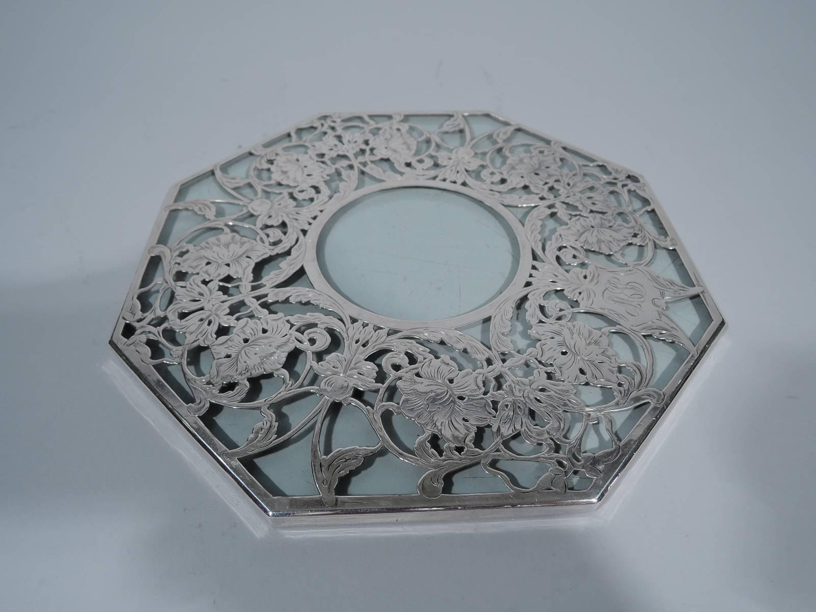 Silver overlay glass trivet. Octagonal clear glass overlaid in silver with scrolled floral garland. Unusual form and lush and fluid ornament. Cartouche has engraved script monogram. Hallmark faint. Condition: Very good with nice patina. Glass has