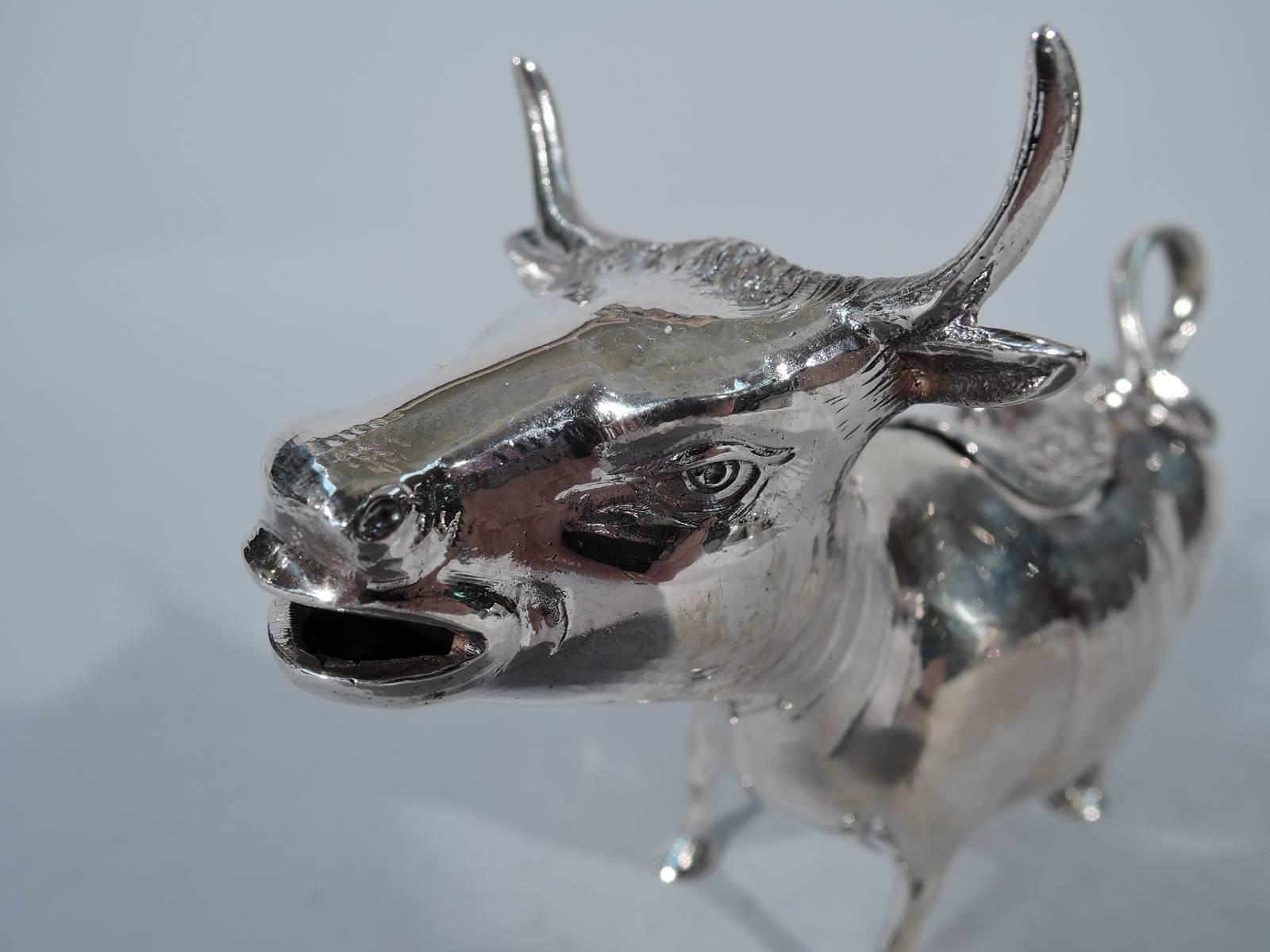 Sterling silver cow creamer. Made in Germany, circa 1950 for Cartier in New York. A sturdy quadruped with flexed ears, sharp horns and ring handle in form of flicked-back tail. Hinged back cover decorated with naif flowers and fly finial. Gaping