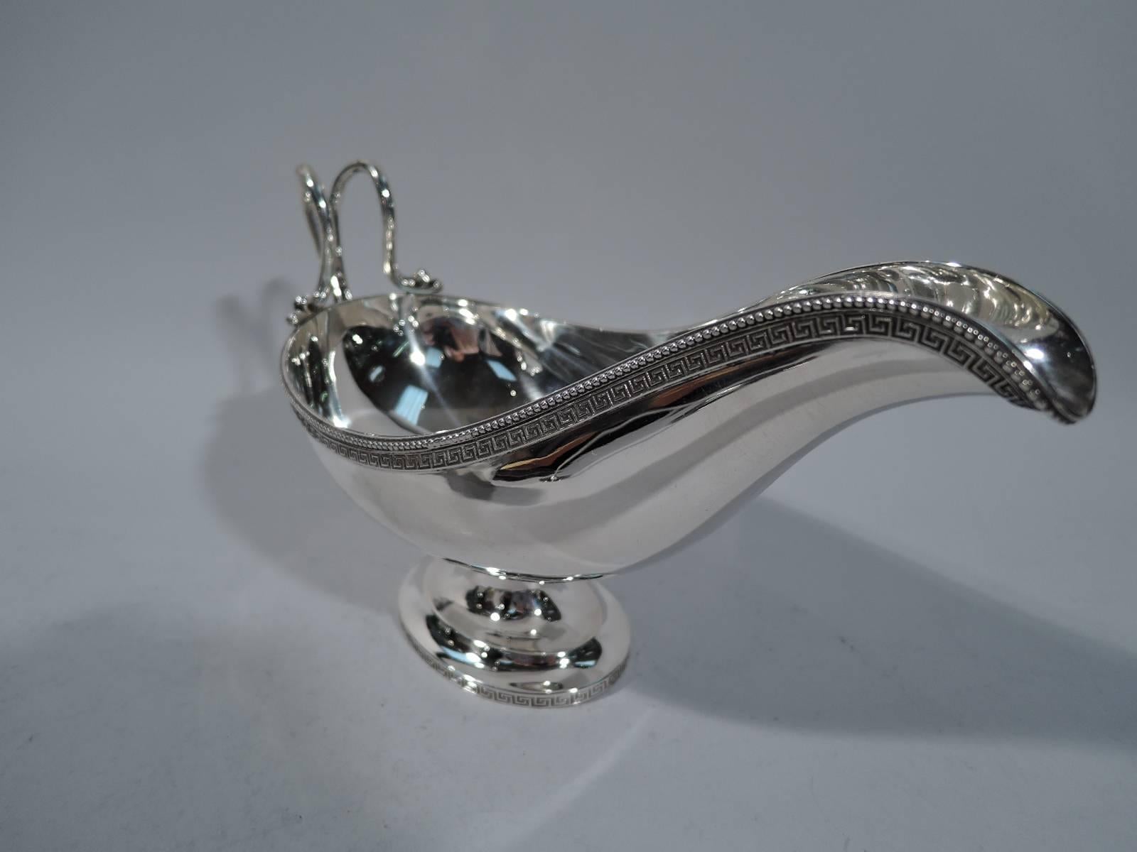 Greek Revival sterling silver gravy boat. Made by Tiffany & Co. in New York, circa 1855. Elongated bowl with helmet mouth, split high-looping handle with leaf mount, short stem, and raised oval foot. Rims have fretwork borders. Mouth rim also has