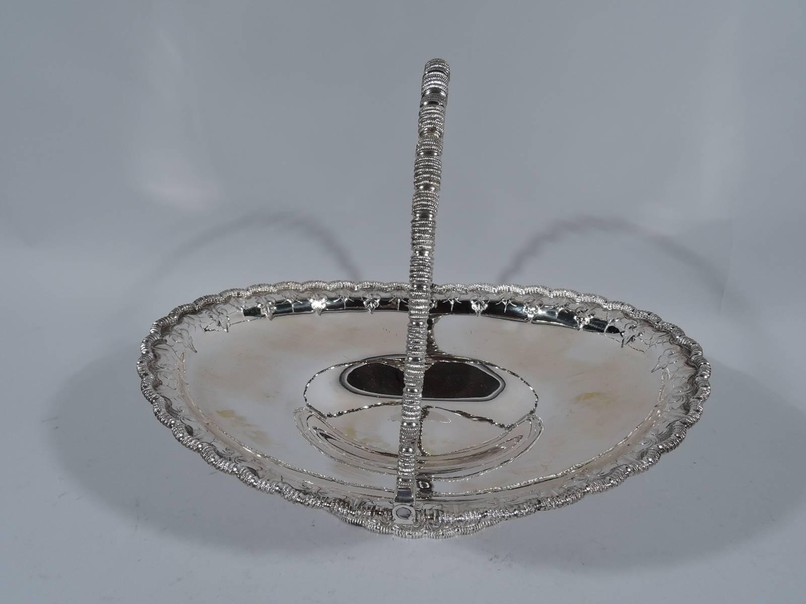 Sterling silver basket. Made by Grosjean & Woodward, 1854-65, for Tiffany & Co., both in New York. Shallow and oval body on raised foot. Rims scalloped and gadrooned as is swing handle. Engraved alternating leaves and fleurs de lis. Early hallmark