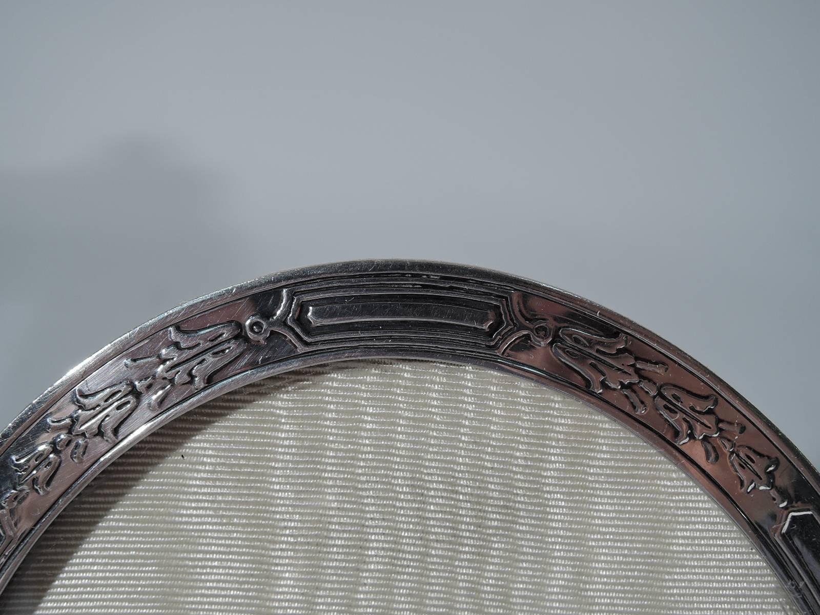 Edwardian sterling silver picture frame. Made by Tiffany & Co. in New York, circa 1910. Oval window and flat border with acid-etched imbricated flowers. Vacant cartouche. With glass, silk lining, and stained wood back and hinged support. Hallmark