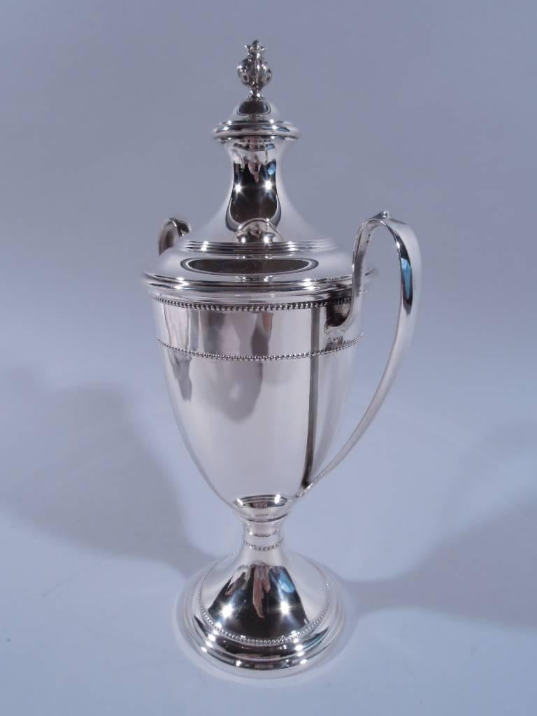 American sterling silver trophy cup. Traditional form with urn body, high-looping capped handles, and raised foot. Double domed cover with bud finial. Beading on body and foot. Reeding on cover. Hallmarked M. Fred Hirsch, a Newark maker active from