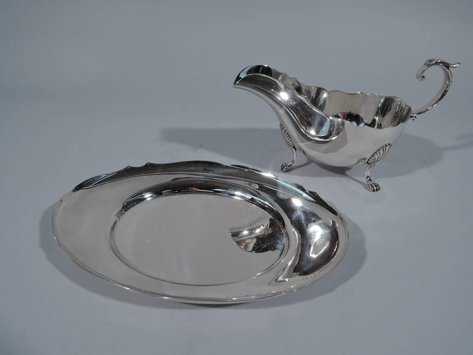 Sterling silver gravy boat on stand in Standish pattern. Made by Gorham in Providence, circa 1920. Boat has scrolled rim, flying double-scroll handle with leaf cap and three paw supports with scallop shell mounts. Stand has oval well and scrolled