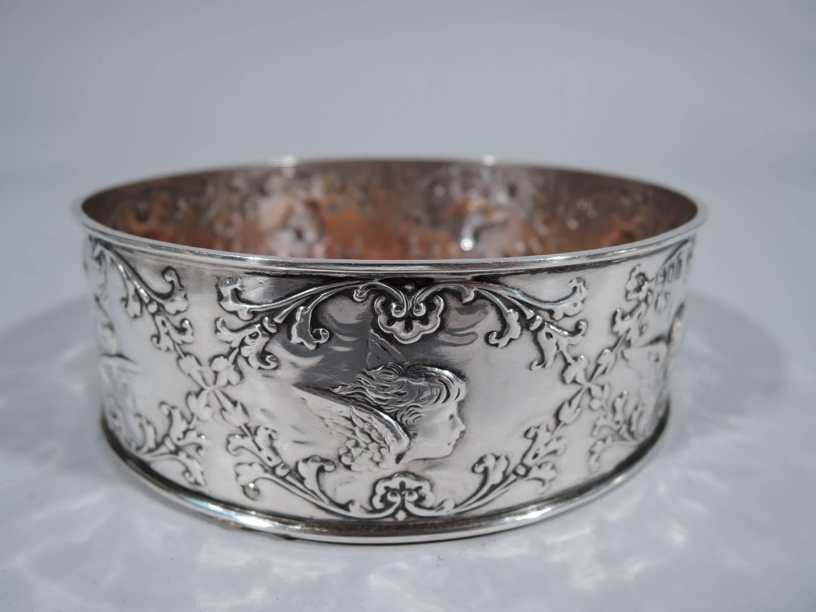 Edwardian sterling silver wine bottle coaster. Made by William Comyns in London in 1903. Straight sides and molded rims. Encircled with floral garland inset with winged cherubs’ heads. Stained-wood well has central silver frame with engraved