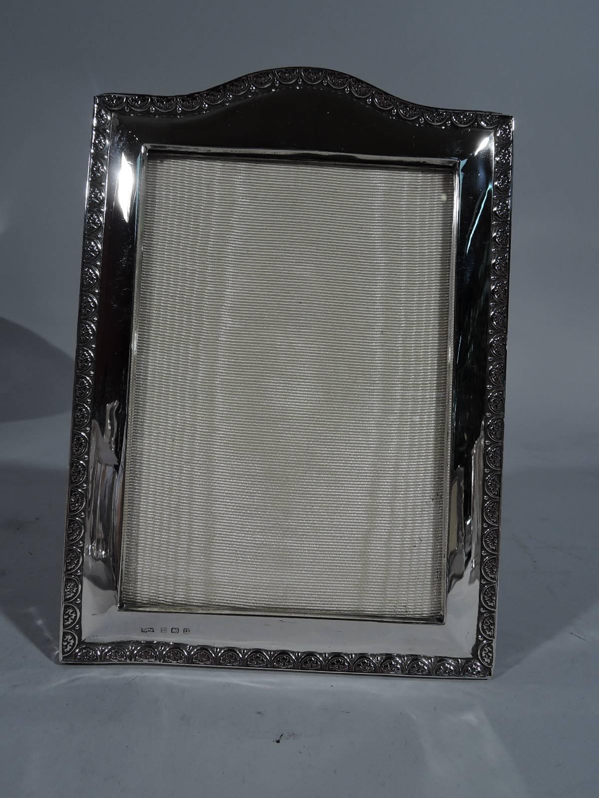 George V sterling silver picture frame. Made in Birmingham in 1913. Rectangular window with tapering border and arched top. Rim has leaf-and-dart ornament and engraved lines. With glass, silk lining, and cloth back and hinged support. Indistinct
