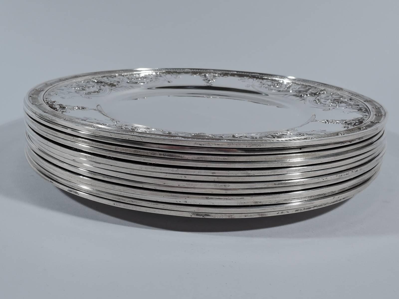 Set of 12 sterling silver dinner plates. Made by Dominic & Haff in New York, circa 1910, for JE Caldwell in Philadelphia. Each: plain well surrounded by neoclassical ornament including paterae, covered vases, and flutes as well as swooping