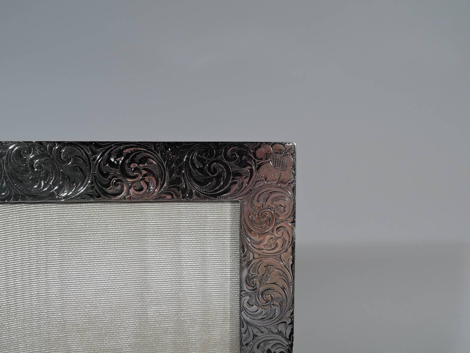 Edwardian sterling silver picture frame. Made by Tiffany & Co. in New York, circa 1910. Rectangular window bordered by engraved and fluid scrollwork and flowers. Tubular cartouche (vacant). With glass, silk lining, and stained-wood back and hinged
