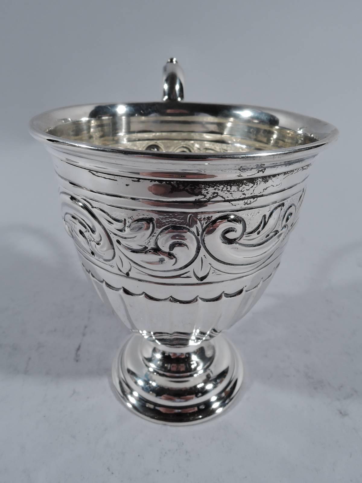 Edwardian sterling silver baby cup. Made in London in 1906. Curved and tapering bowl with half fluting and scrollwork band. Capped s-scroll handle and stepped foot. Indistinct maker’s mark. Measures: Weight: 4 troy ounces.