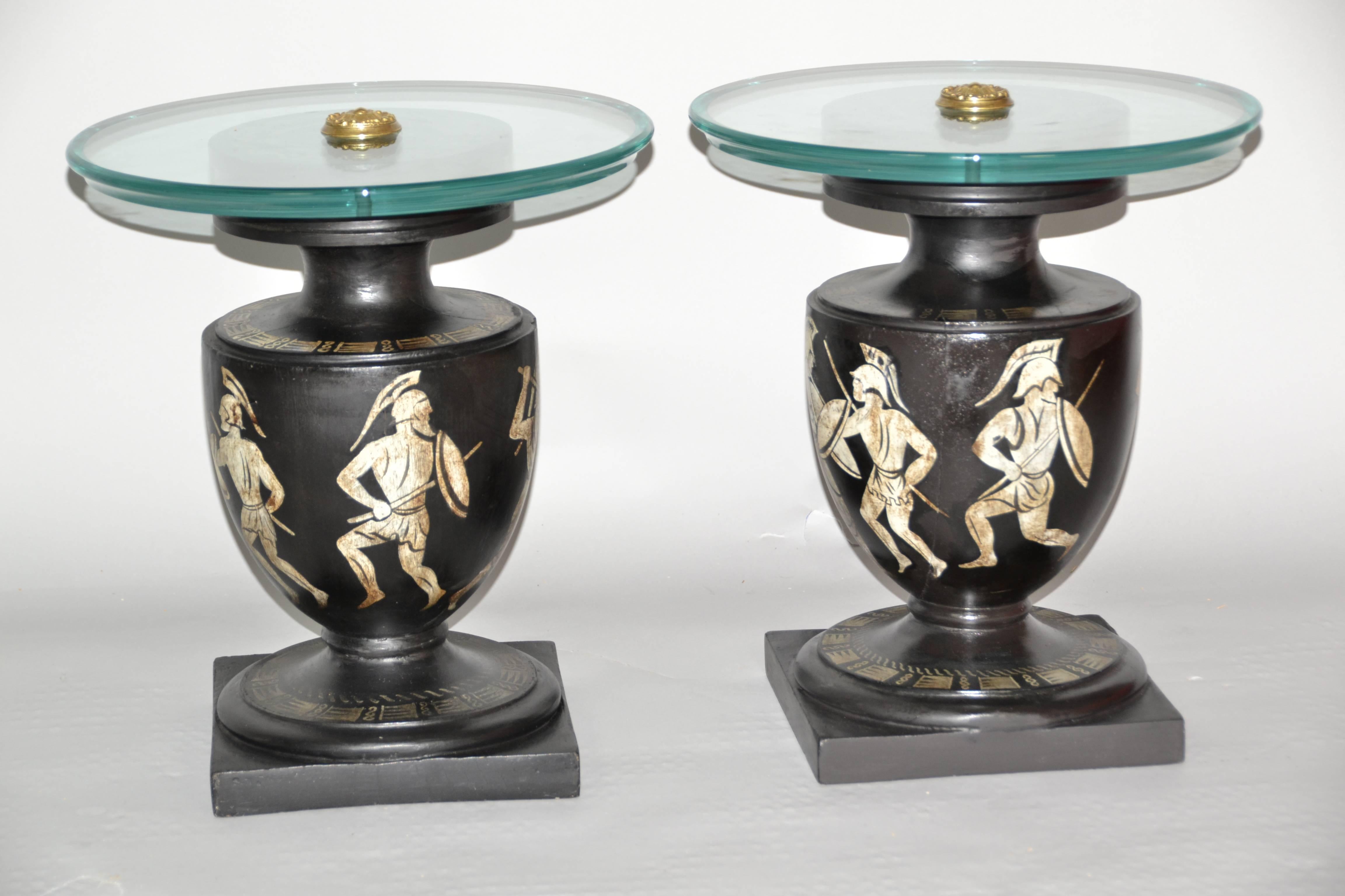 A Neoclassical pair of mid century Italian black and white painted wooden urn based side tables with aquamarine glass tops. The 1940's end tables with hand-painted white gladiators on black background decoration are based on classical Greek pottery