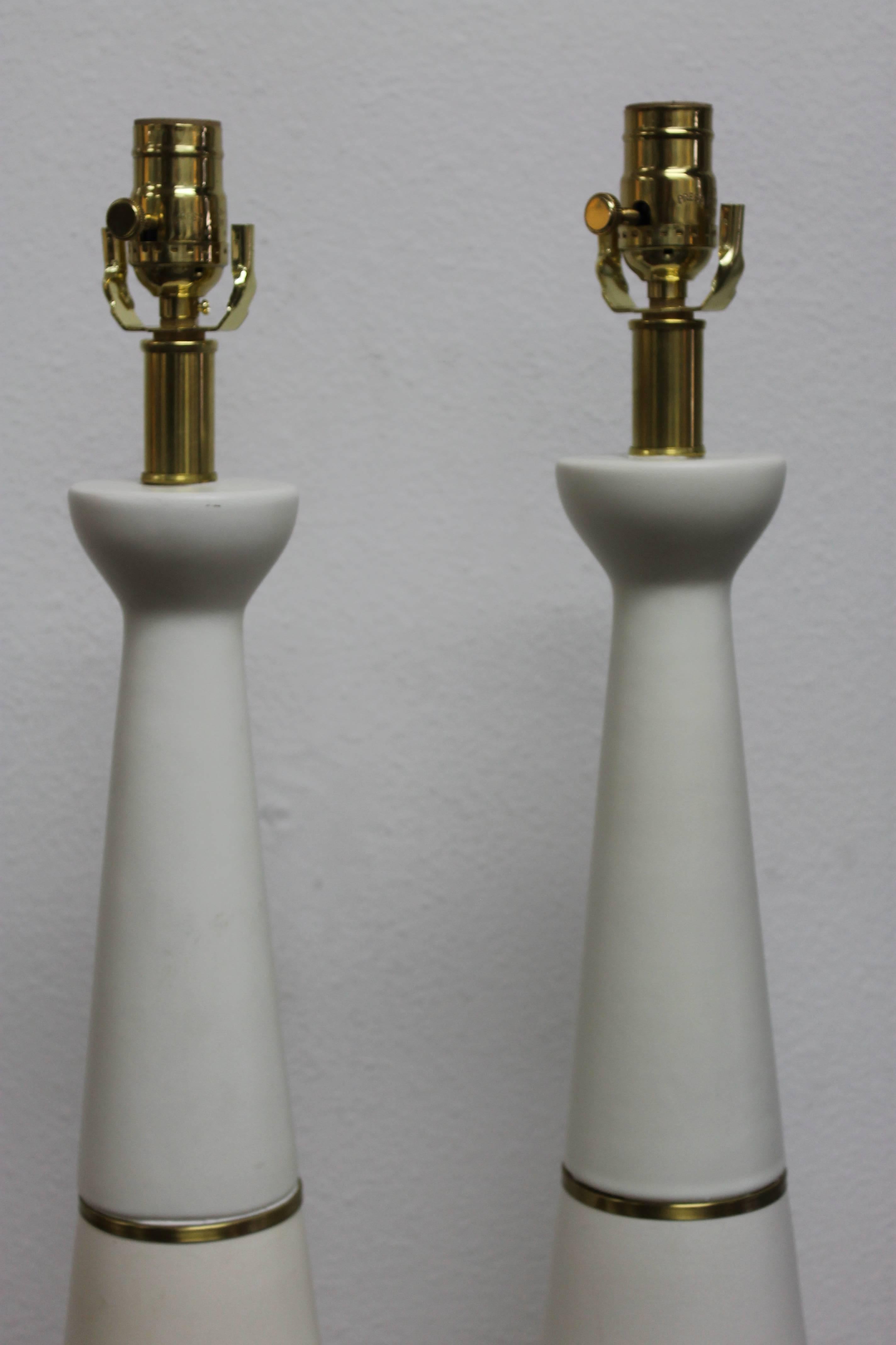 Pair of white ceramic lamps with brass rings. Measures: Wood base is 6.5" diameter. Ceramic portion is 25" high. Total height from base to the bottom of socket is 28".  Lamp shades not included.
  