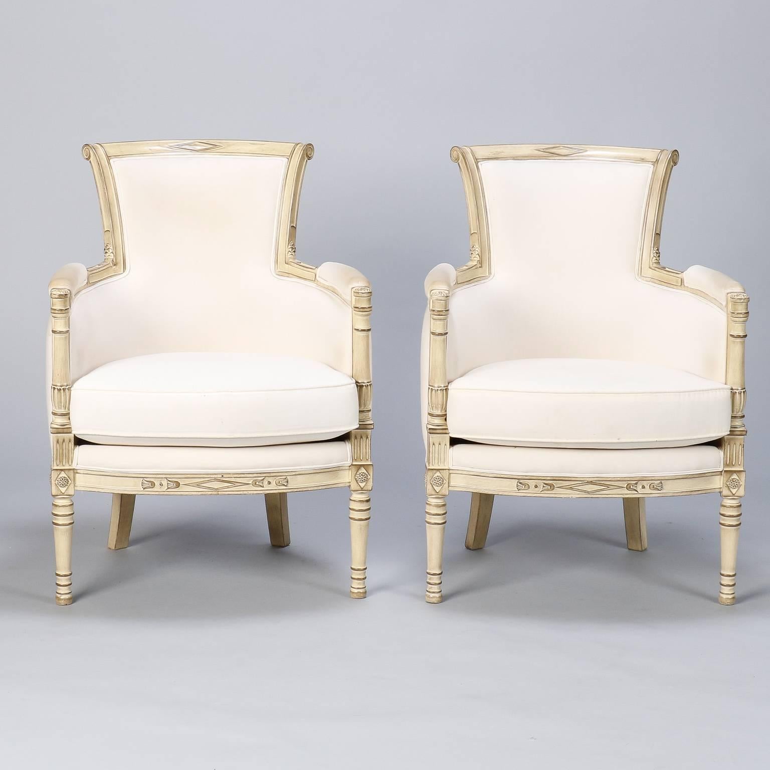 Mid-Century pair of Louis XVI style bergeres with carved and antique white painted frames. Chairs have curved base, narrow, flared back rests, padded arms and carved details. Original upholstery has been stripped down to the under liner muslin and