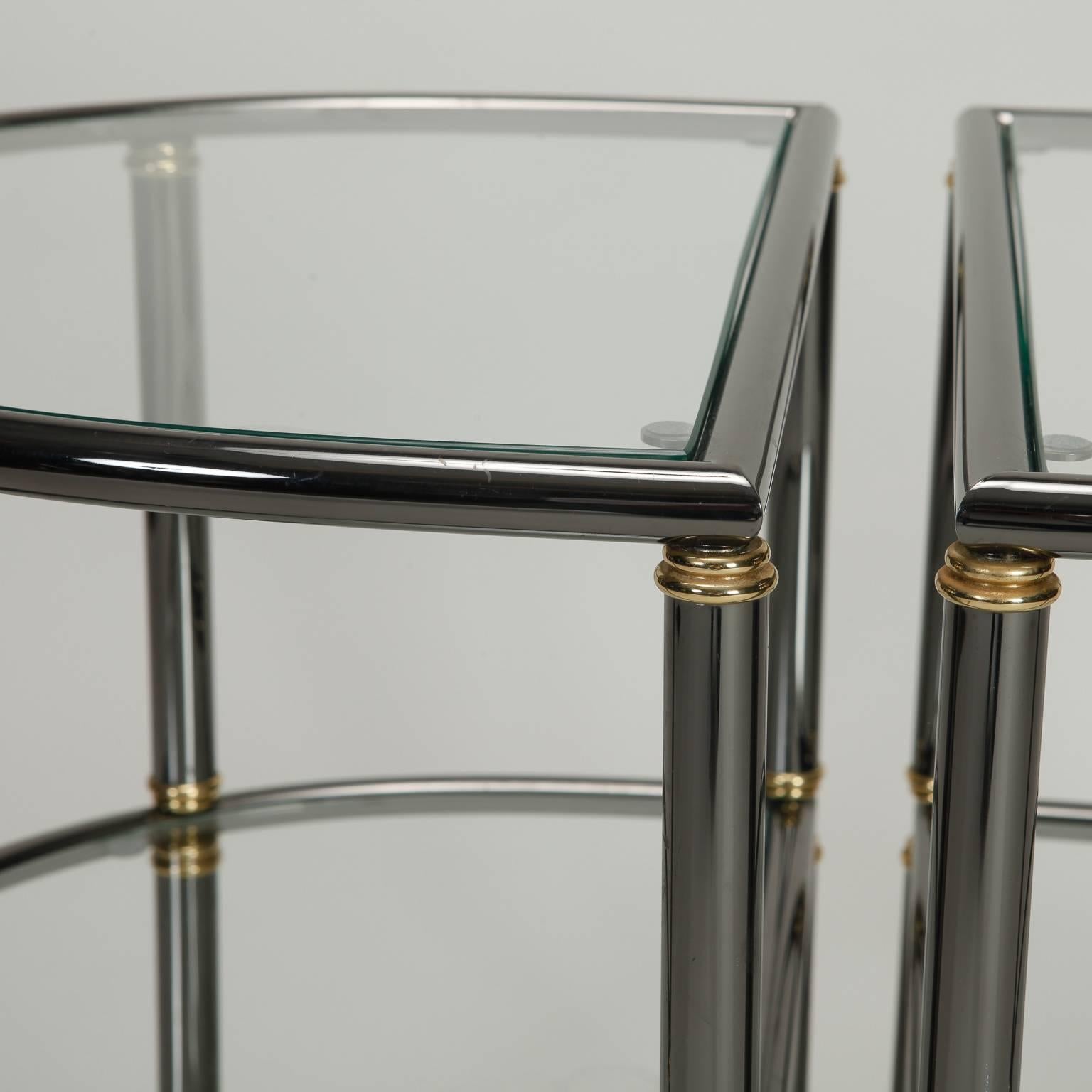 Paire de tables d'appoint demilune à deux niveaux avec des cadres tubulaires en métal gun, des accents en laiton et des étagères en verre, vers les années 1970. Vendu et tarifé comme une paire.
 