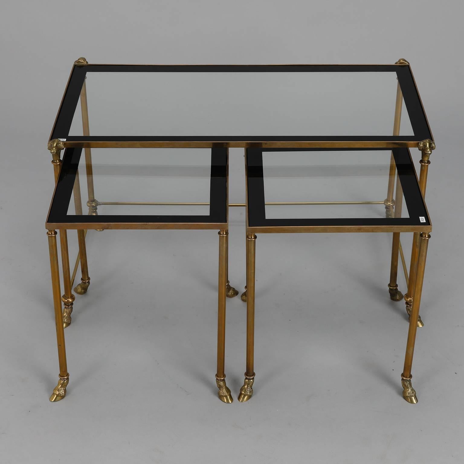 Mid-Century set of three French brass frame and glass nesting tables. Pair of two smaller tables nest side by side under larger table. The larger table has ram's heads at tops of legs. All tables have hoof form brass feet and black enamel framing