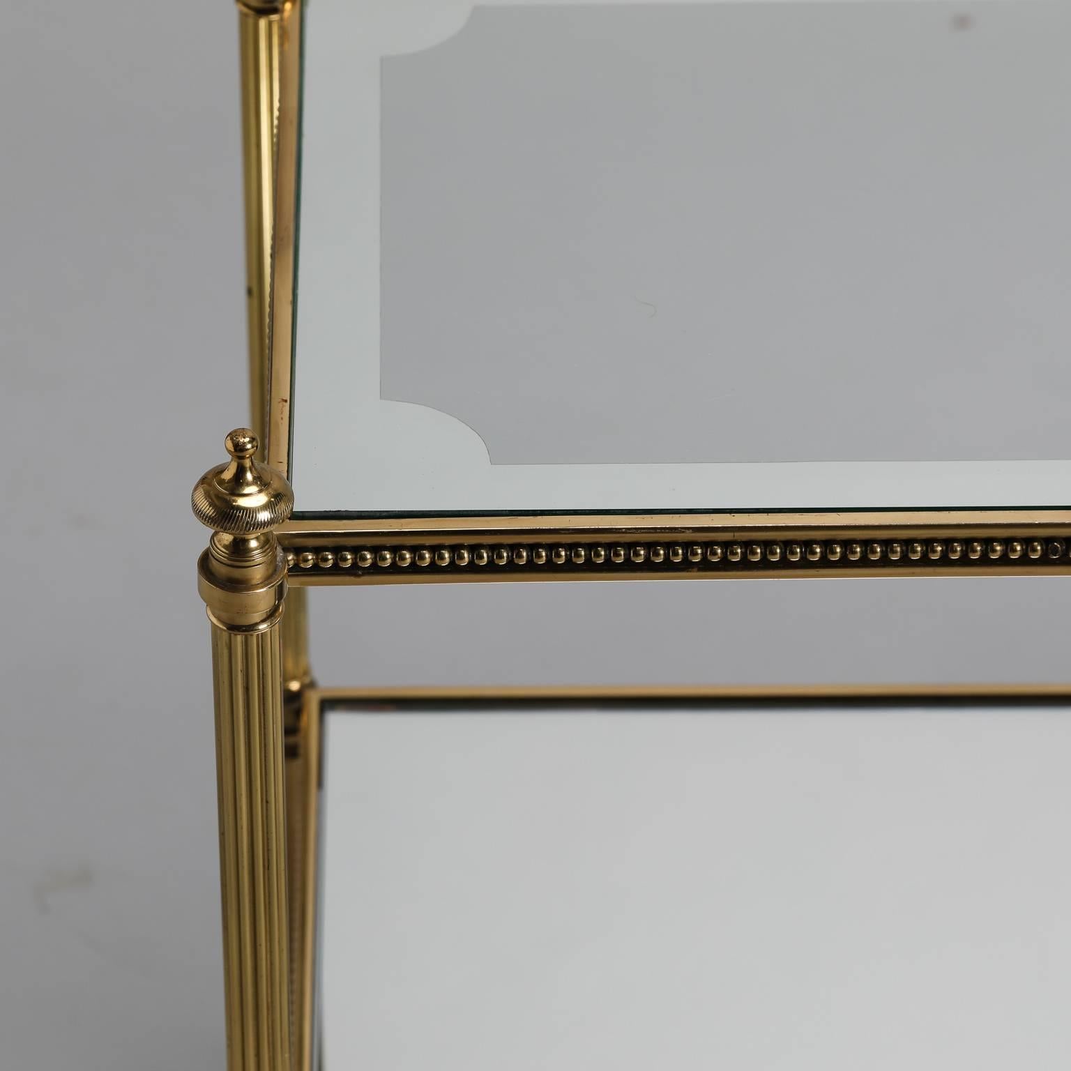 Mid-Century pair of Italian brass, glass and mirrored glass side tables date from the 1960s with neoclassical styling. Two-tier square tables have fluted brass legs, decorative finials and beading and glass table tops with wide, decorative dark