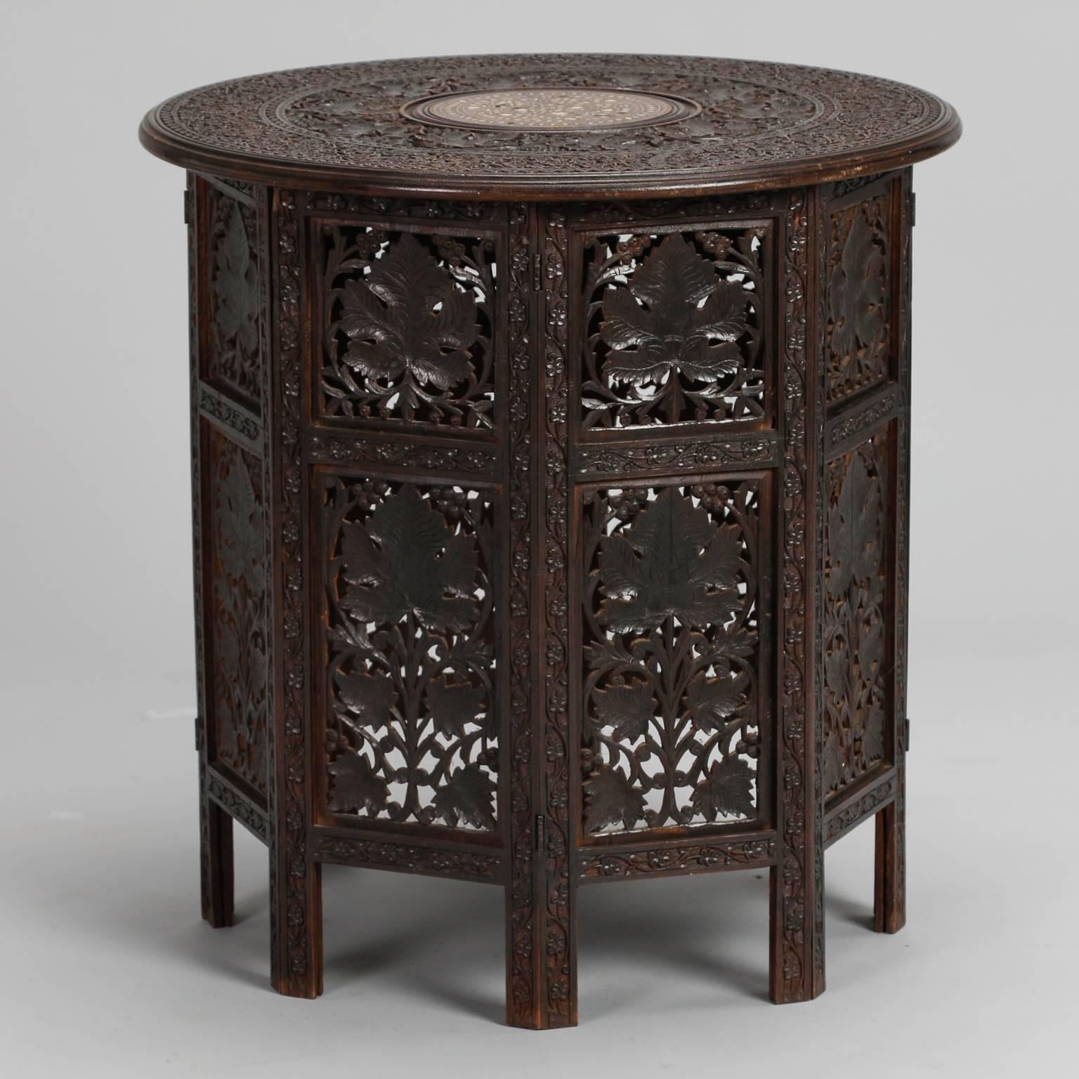 Dark wood side table is intricately carved with a design of leaves and vines, circa 1930s. Round top is removable and the base is made up of hinged panels that can be folded flat. Center of tabletop has a design of inlaid bone.
