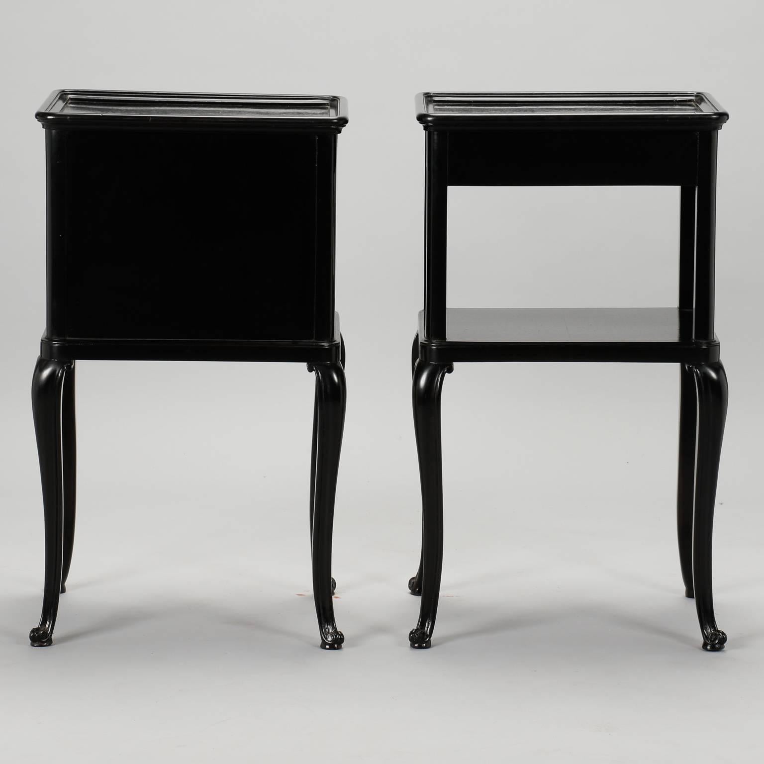 Pair of circa 1930s slender Art Deco side tables found in France with new ebonized finish. Side tables are the same size with slender cabriole legs, silver tone hardware. One table has a hinged cabinet and drawer while the other has a lower shelf,