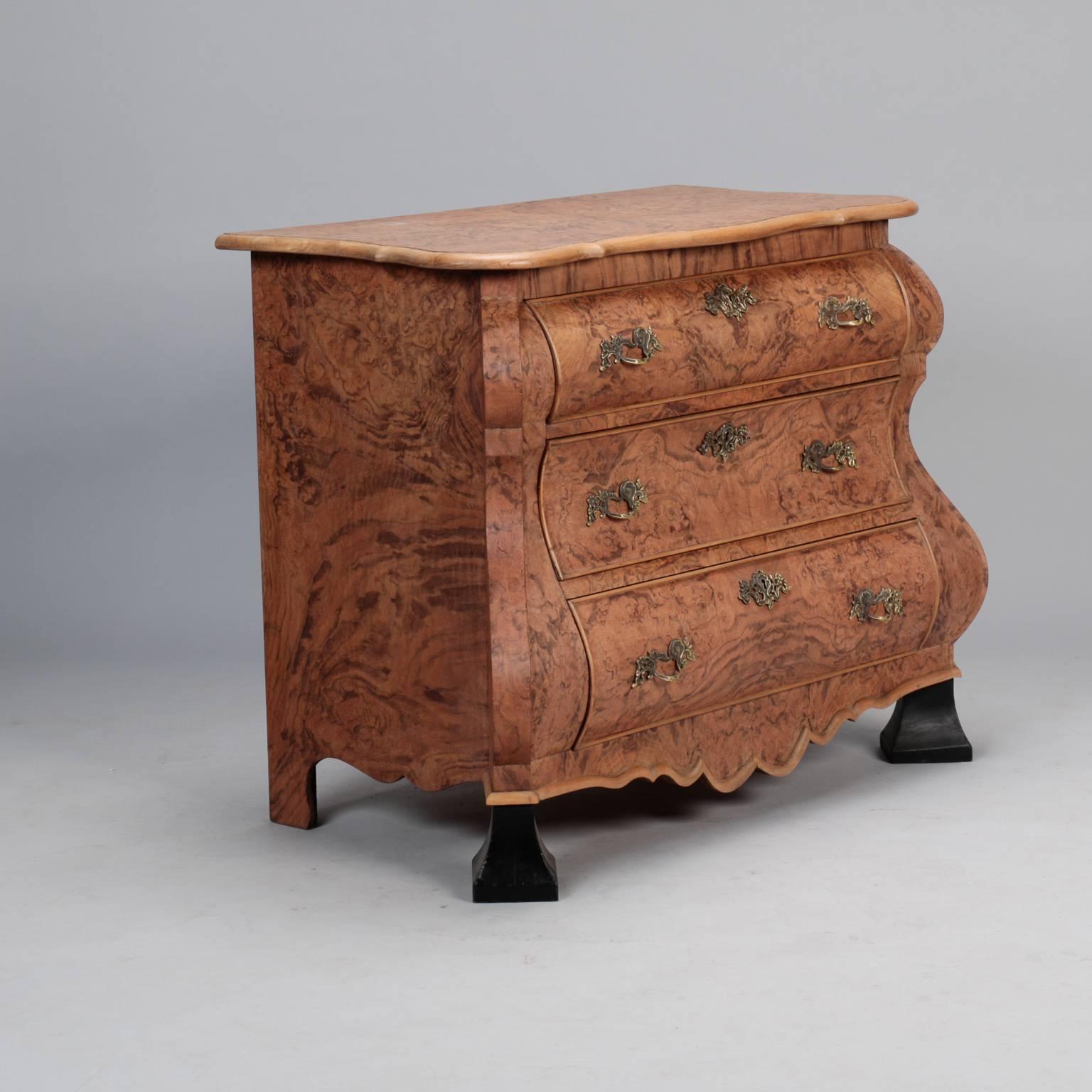 Circa 1920s bombe chest has burr walnut exterior, three locking drawers, antique brass hardware, wide ebonised feet and a curvy apron. 

