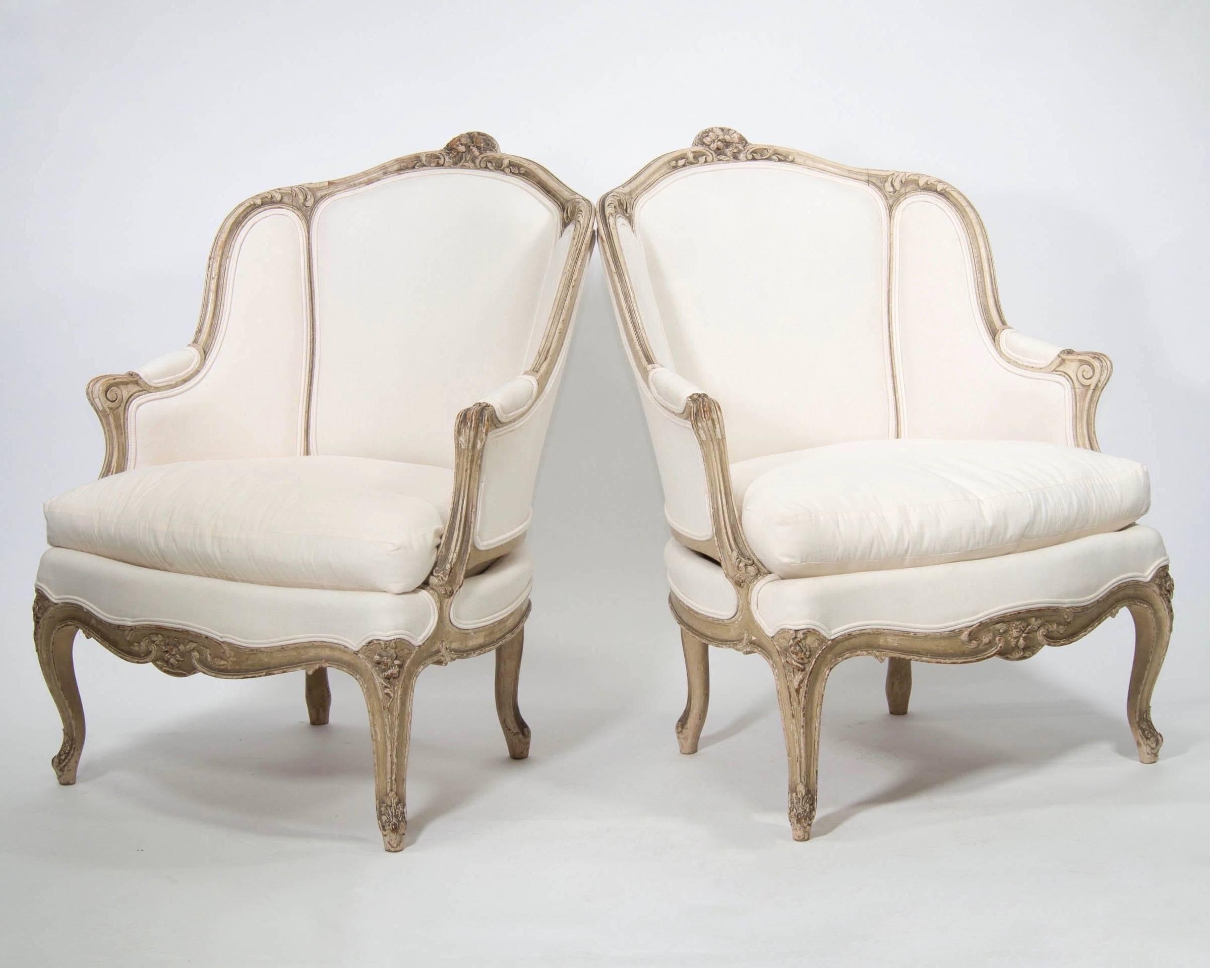 Pair of bergeres feature ecru color painted frames with carved details on crest, apron, arms and legs, circa 1890s. Wingback style sides, padded armrests, down filled seat cushions and cabriole legs. Upholstered in muslin, these chairs can be