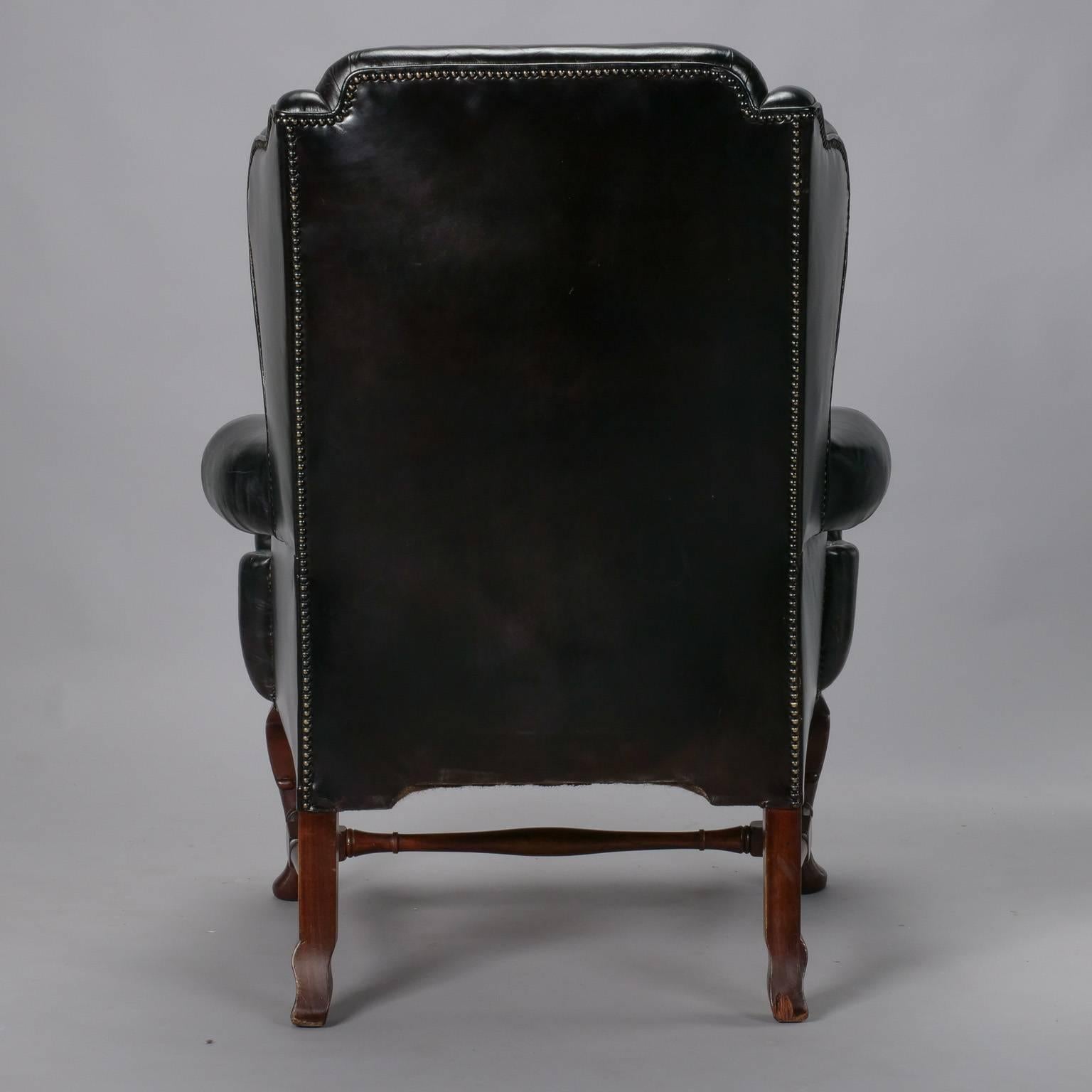 Traditional English wing chair features generous proportions, black leather upholstery with brass nailhead trim, high back, rolled arms and dark wood legs and stretcher, circa 1940s.
 