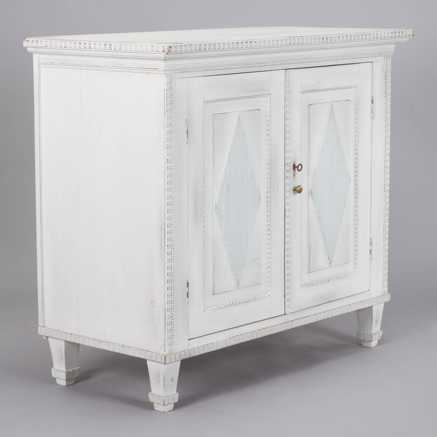 Swedish cabinet has two hinged doors, a single internal shelf and white painted finish with subtle contrast on carved diamond detail, circa 1900. Carved details along edges and legs, working key lock.