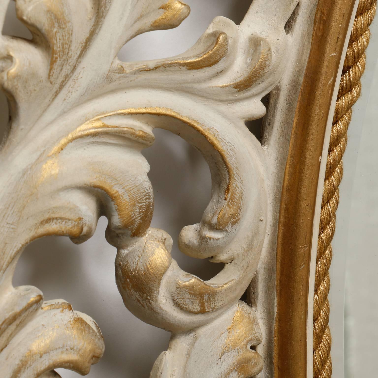 Italian bevelled edge oval mirror is has an open work carved and painted frame of leaves, circa 1920s. Antique white finish with gilded edges.  Actual Mirror Size:  24.25” h x 19.25” w
      