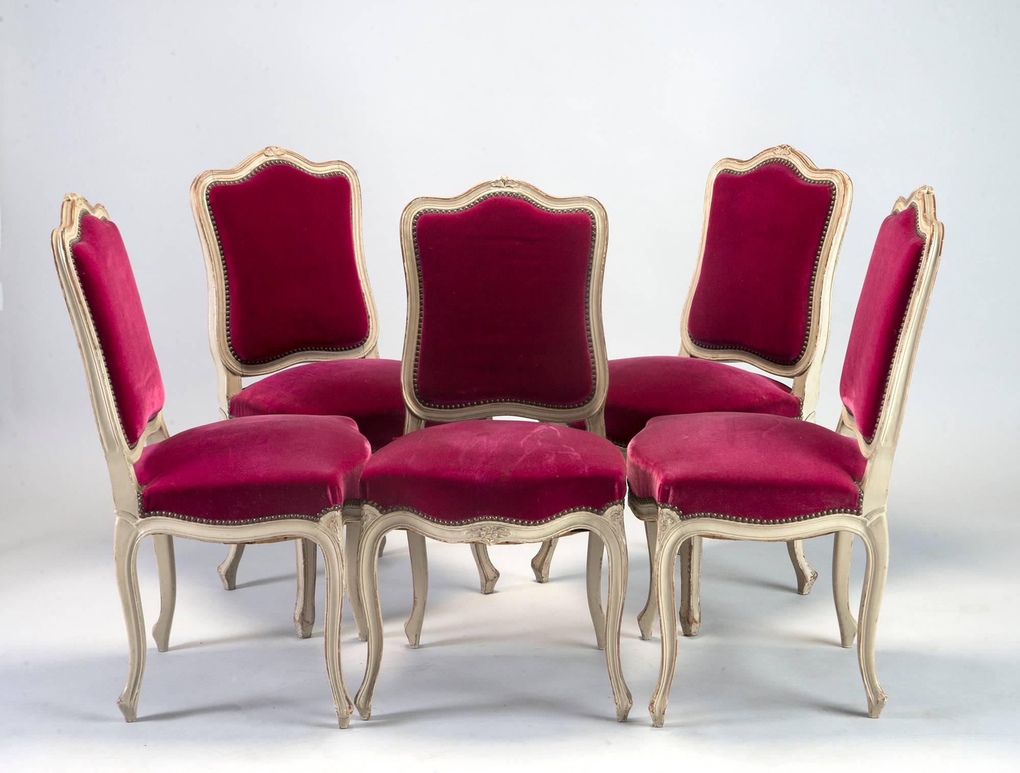 Set of 12 circa 1920s French dining chairs with camel back seat rest, antique white painted frames with carved floral crest and carved floral medallions at the knees. Older magenta velvet upholstery is presentable but shows some wear, slight soiling