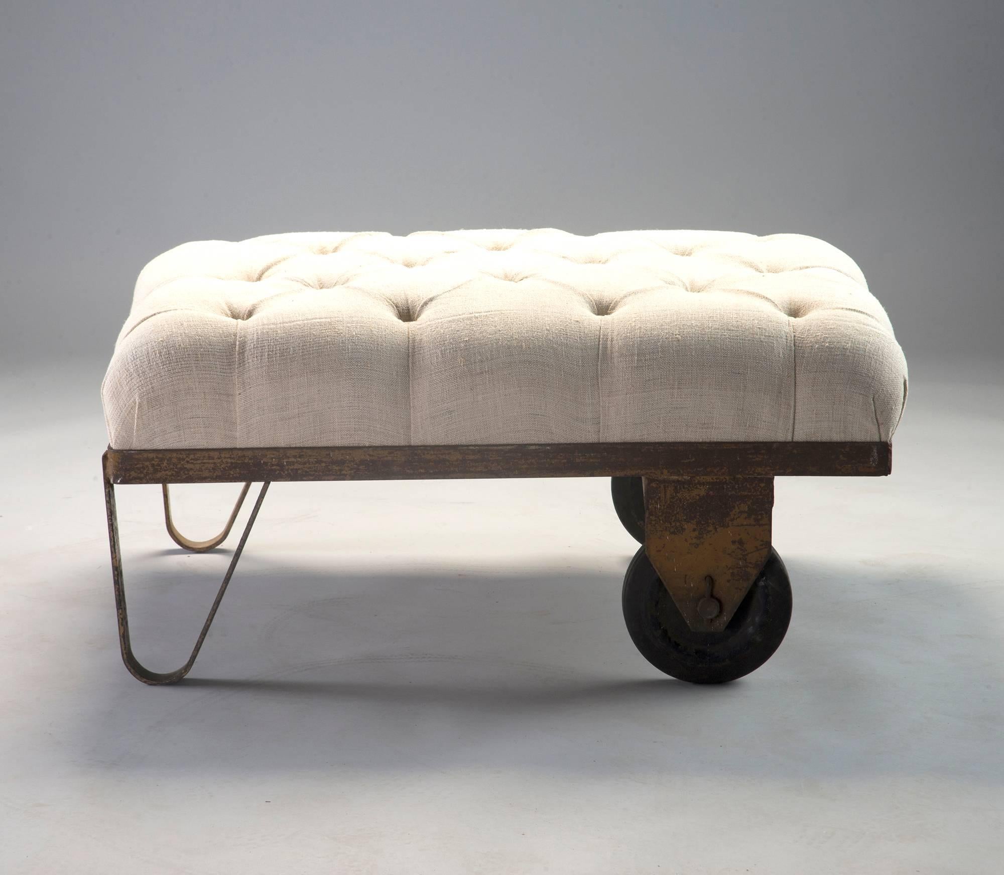 Stool, ottoman or bench made with circa 1930s industrial factory wheelbarrow base. Base of cast metal has remnants of original painted finish and rusty patina. Two rubber wheels and legs with newly upholstered top in tufted, natural colored linen.