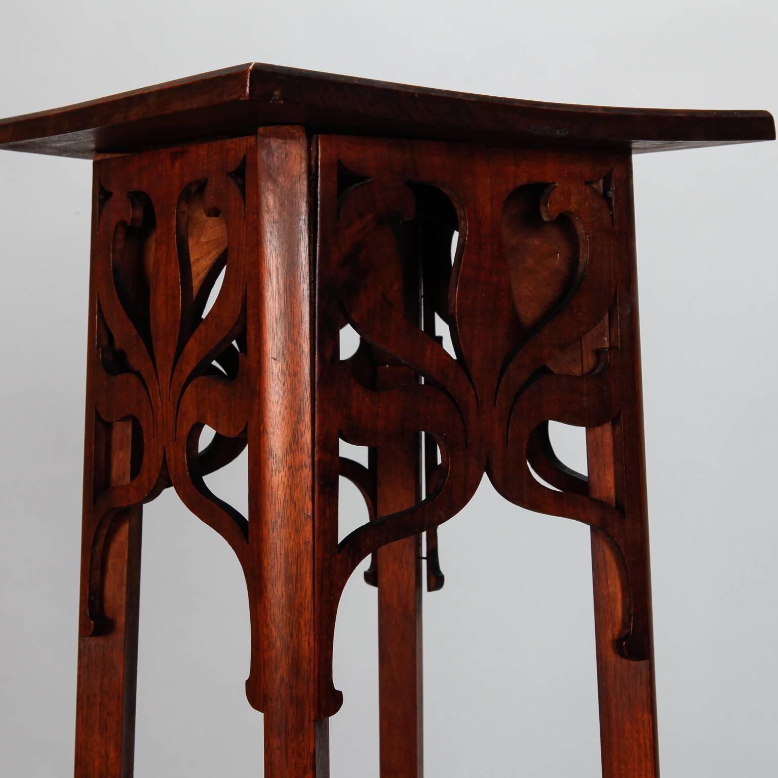 Tall narrow American wood table for displaying a statue or plant. Table features classic Arts and Crafts style with decorative, open work apron, and details carved in relief on table top and lower tier. Table top measures 11.5
