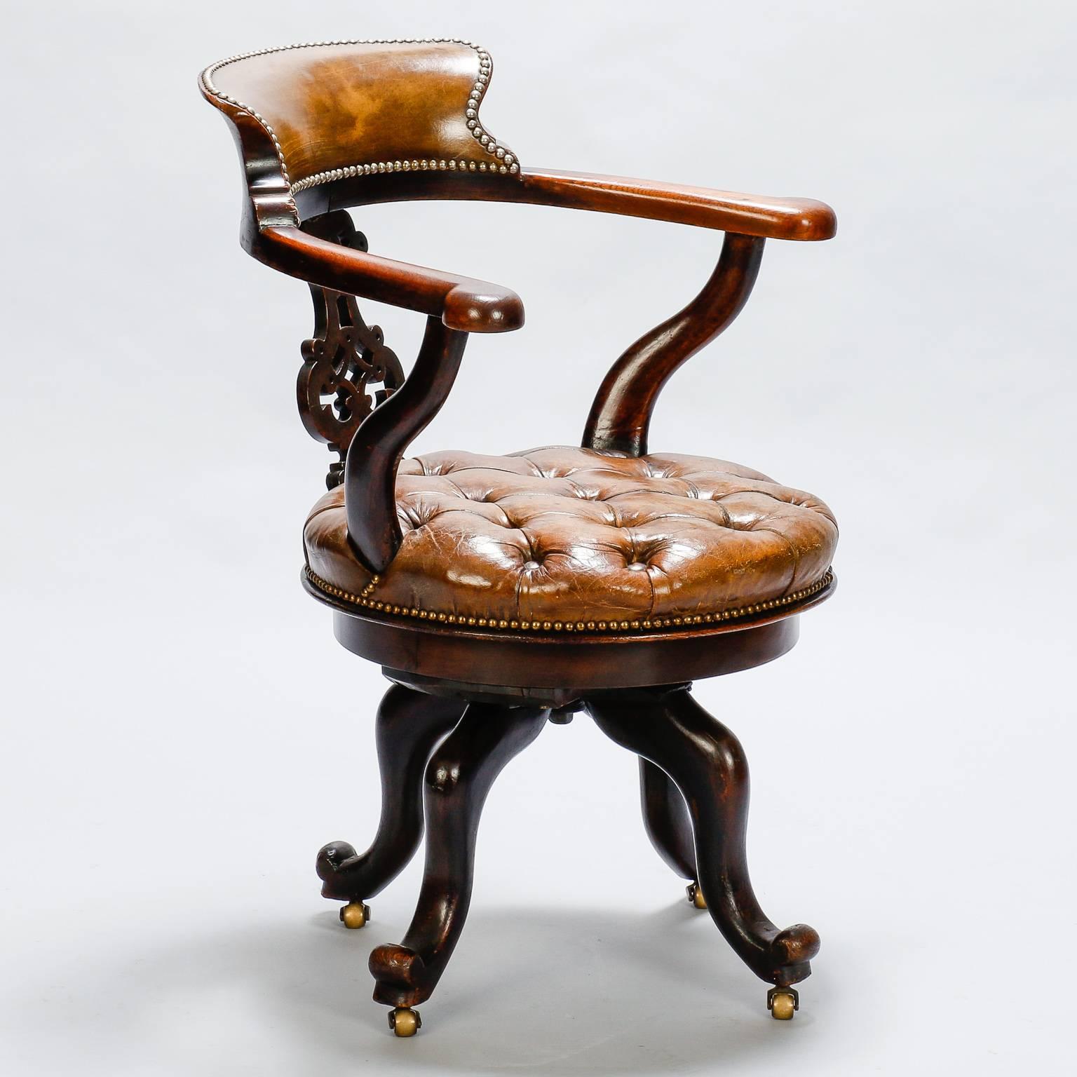Circa 1920s French desk chair has mahogany frame with four cabriole legs on brass casters, a tufted leather seat that swivels, arms, a decorative carved back splat and padded leather back rest with brass nailheads. Seat is 19” high and 19” deep.