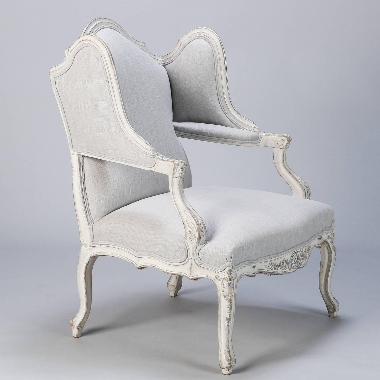 Circa 1940s bergere has Regency style frame with antique white painted finish. Winged sides meet the arms, the chair has a crested back rest,  cabriole legs and apron with carved details. Newly upholstered in pale gray linen fabric. 

Arm Height: 