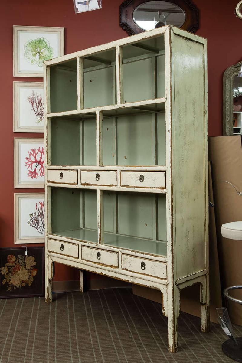 Chinese cabinet has a cream color lacquer finish on the outside with a contrasting celadon interior, circa 1900s. The top level features three open shelves over two larger shelf compartments framed with narrow functional drawers. Brass ring pulls.