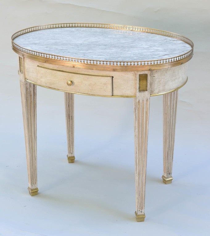 End table, having a painted finish with natural wear. its oval top of white marble with gray veining, surrounded by a pierced gallery of polished brass, over brass-trimmed apron with single frieze drawer, raised on square-section, fluted, tapering