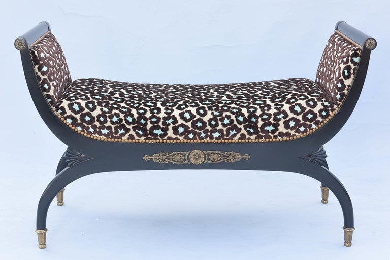Neoclassical style curule bench, by John Widdicomb, having a black lacquer frame, its upscrolling, padded arms finished by rosette, continuing to an apron decorated with bronze ormolu of cast anthemion and palmettes, upholstered in leopard chenille