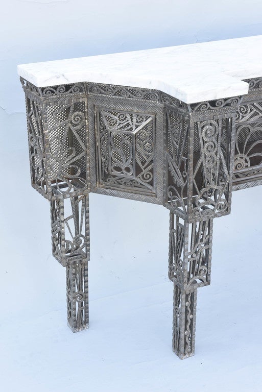 Extraordinary Art Deco console, having a breakfront top, with canted sides, of Carrara marble, on a conforming table base, its hand-hammered steel frame inset with scrollwork and foliate motifs, the apron having double doors, raised on