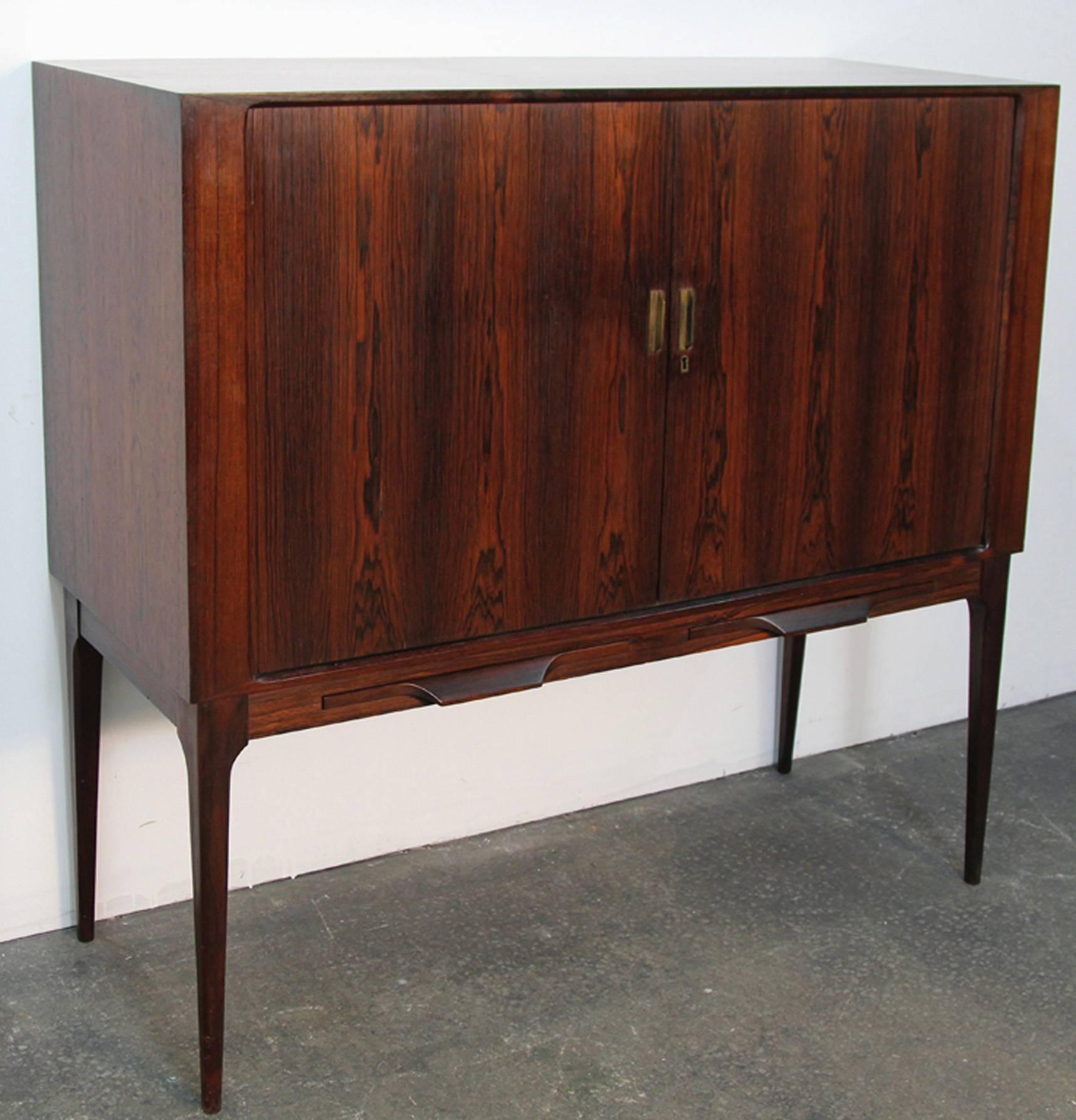 Beautiful rosewood bar cabinet designed by Kurt Ostervig featuring stunning tambour
doors, two interior black drawers, and dual pull-out drink preparation trays.