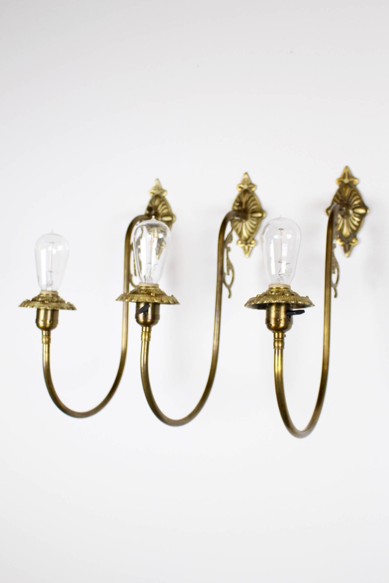 This set of three Victorian style brass wall scones were converted from gas to electricity, circa 1910. They are fitted with bare tungsten bulbs and implore a sense of quirk and whimsy with their airy Silhouette. These sconces bring instant