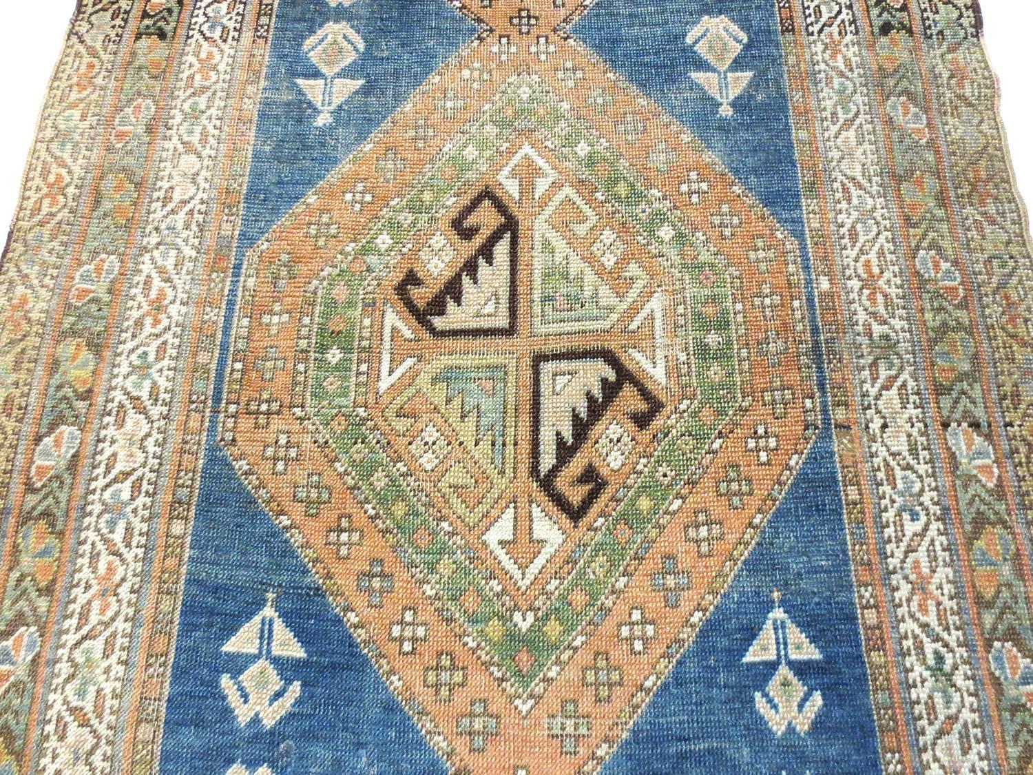This is an antique Kurdish runner, circa 1900s. It features bold geometric medallions dominating on blue field and use of symbolic tribal motifs. Shades of peach, browns, greens, yellows ivory border provide a vibrant array of colors while multiple
