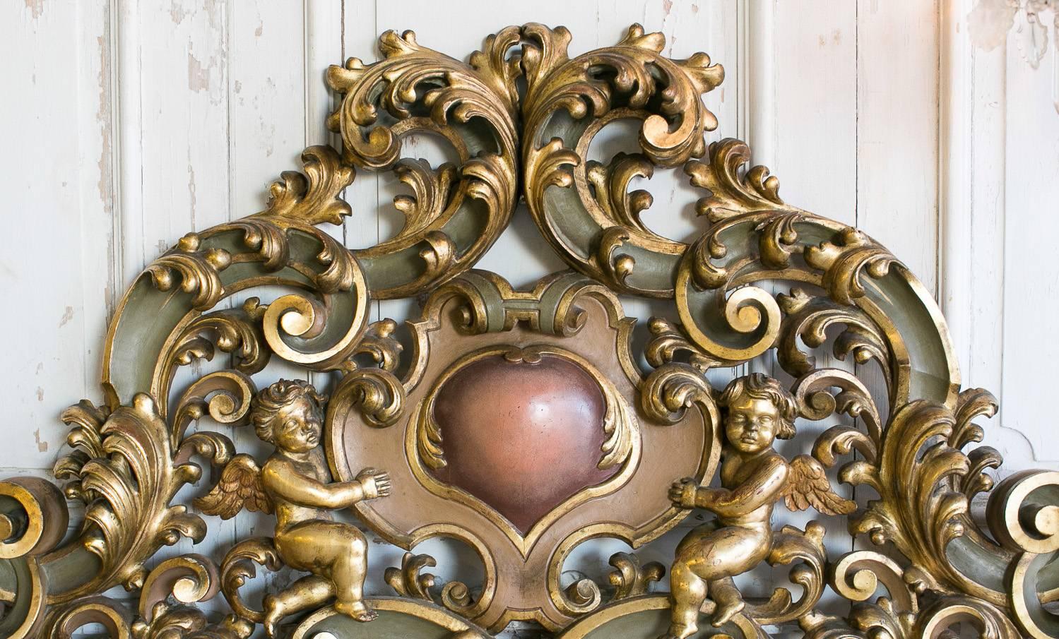 Elaborately carved Baroque-style headboard painted gilt with dark olive contrasts. This opulent queen size headboard features a heart-shaped centerpiece held by two cherubs. Stunning leaf carvings swirl and curve throughout this ornate, Italian-made