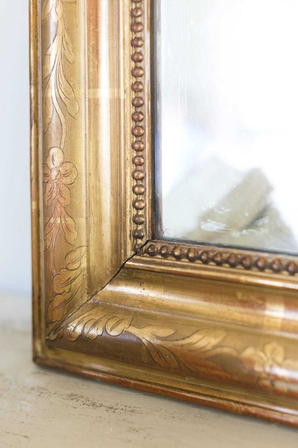 This Louis Philippe mirror features simple floral motif buttons where the glass connects. The gorgeous piece in muted gilt rubs back to umber red in the distressed frame decorated with subtle etchings. Light chipping in the corners and the