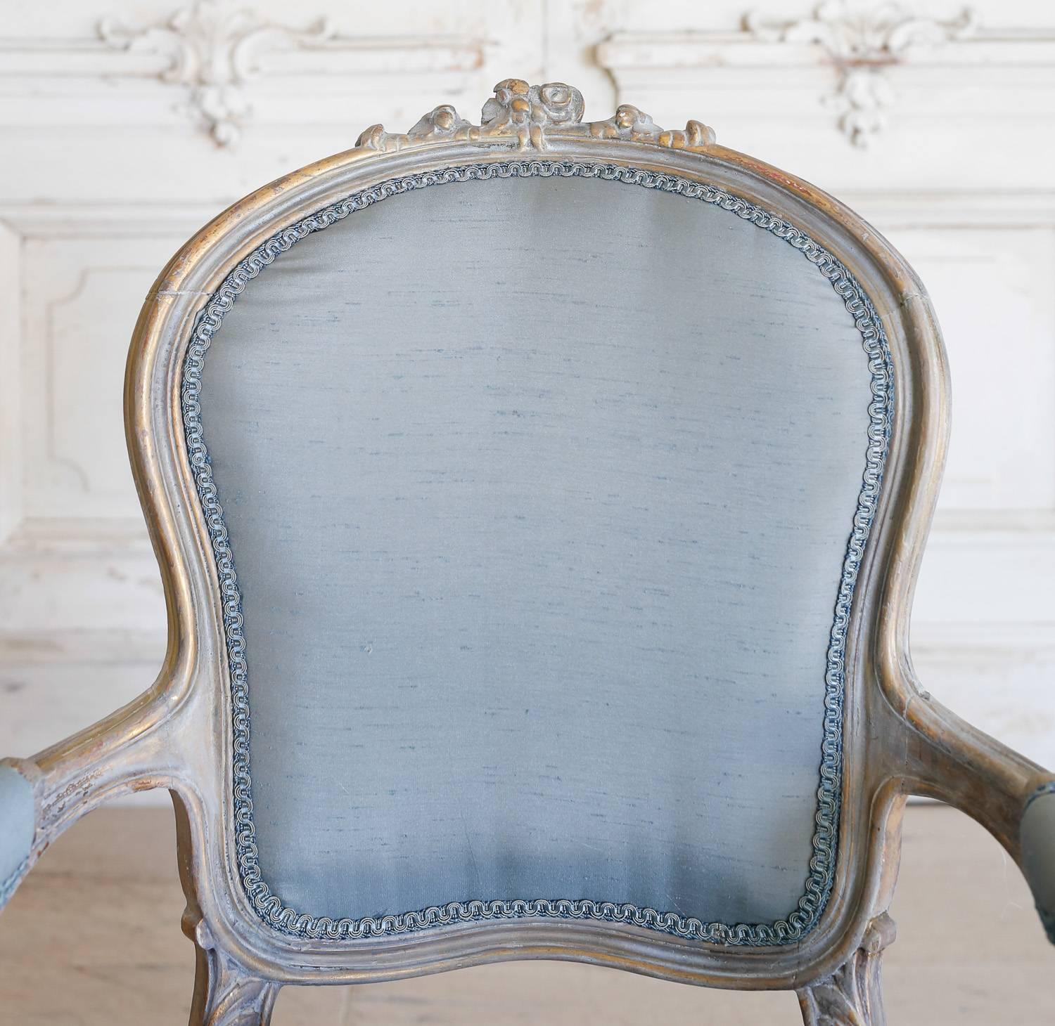 Gorgeous pair of vintage armchairs upholstered in blue silk and finished in a white washed, distressed wood frame. Two rosebuds adorn the crest and scroll down to a carved, floral apron. The Louis XV legs offer a comfortable balance while keeping