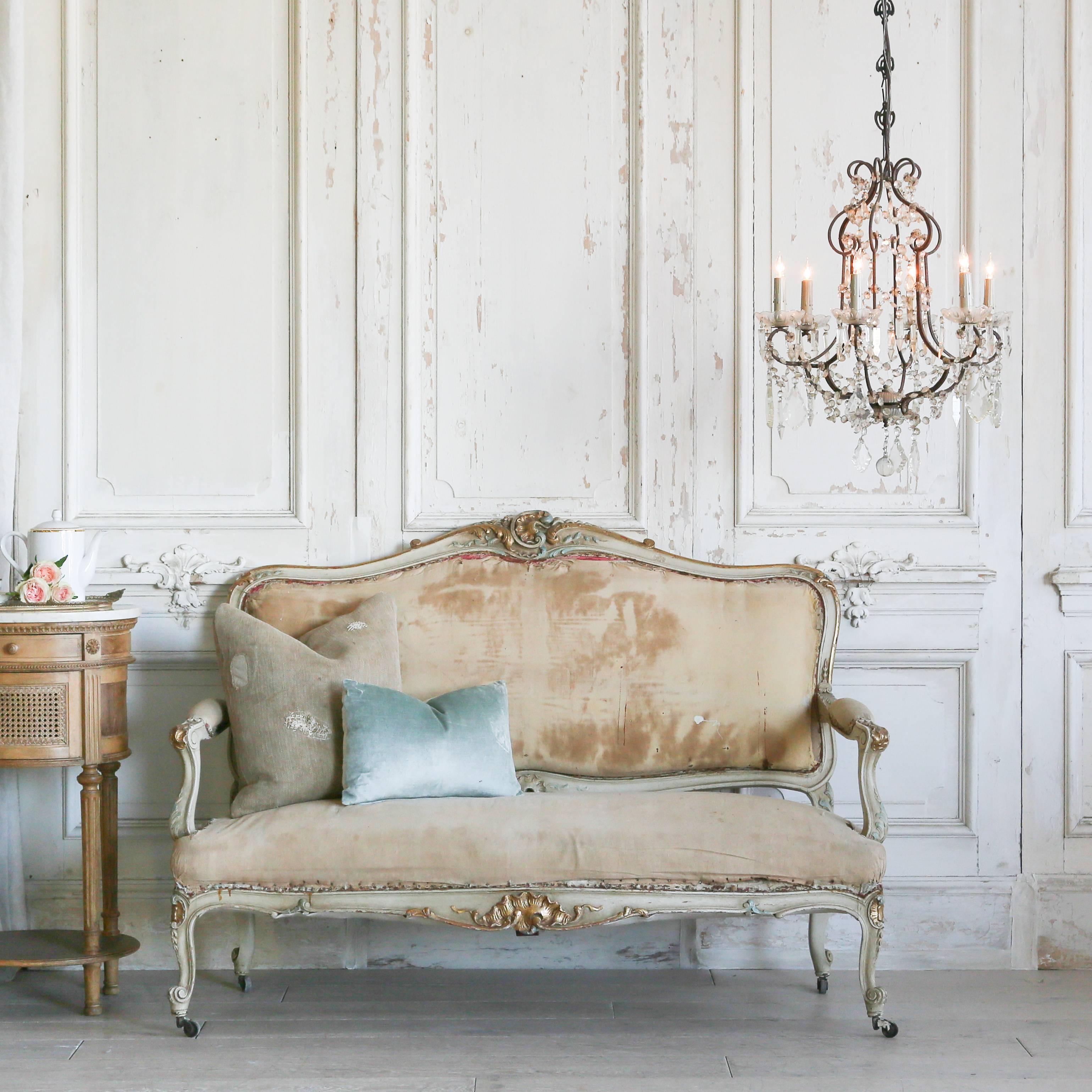 Darling antique settee in distressed bone, gilt, and turquoise tones. This sweet love seat has a prominent crest with swirl and ocean motifs that gleam golden against the blue and neutral paint. The classic Louis XV legs sit atop small rollers.