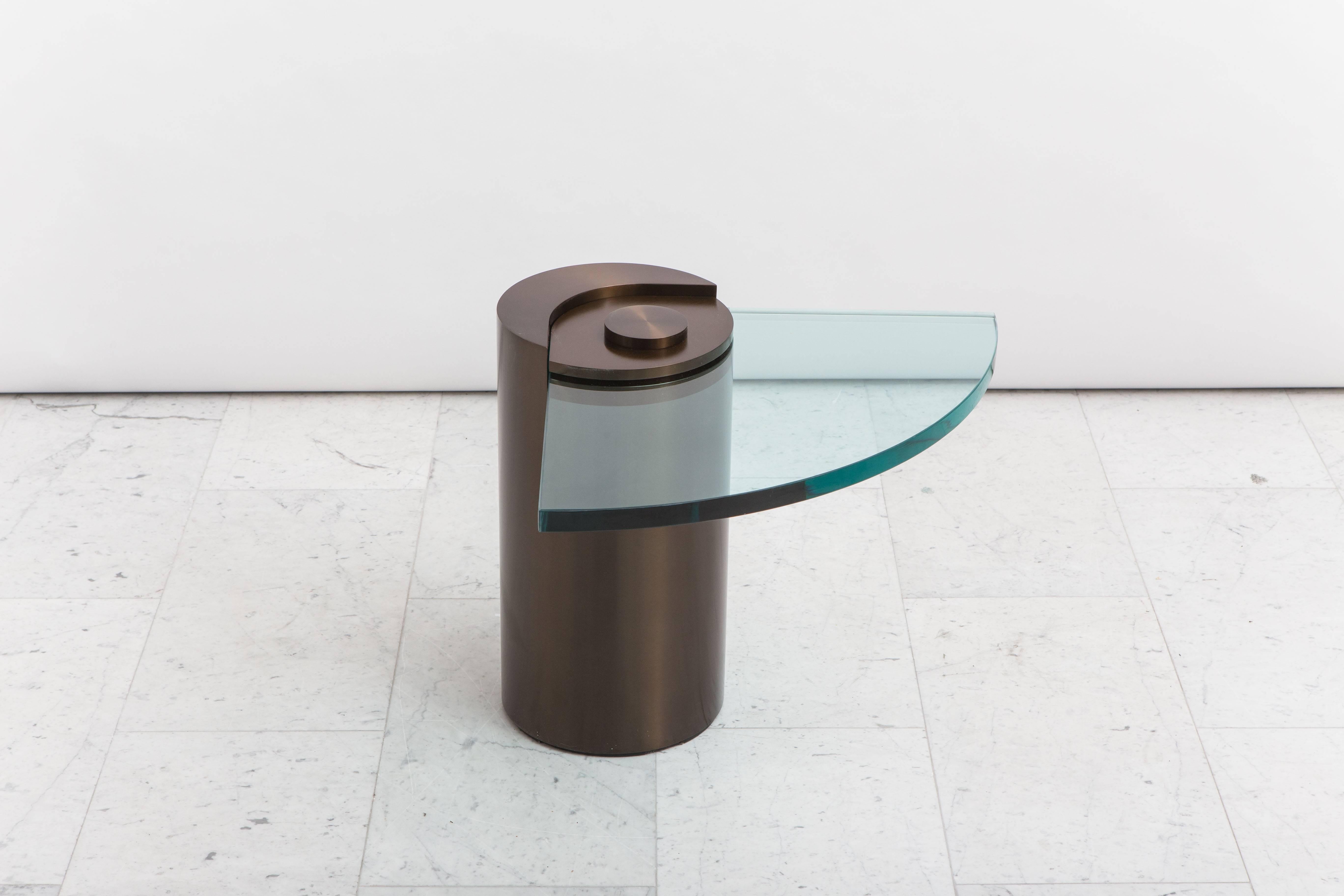 An iconic design by Karl Springer, the unique sculpture leg table debuted in 1971 and is being reissued today exclusively with Todd Merrill Studio. Featuring a cylindrical metal base in royal bronze, an elegant cantilevered fan-shaped glass extends