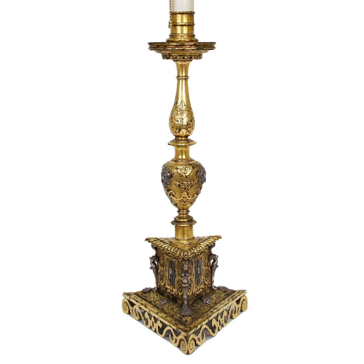 Inspired by the designs of 16th century Italy, this antique, candlestick lamp has a candle-cup and circular drip pan on a baluster turned standard and is raised on a stepped triangular base with a conforming pierced plinth. It is cast throughout