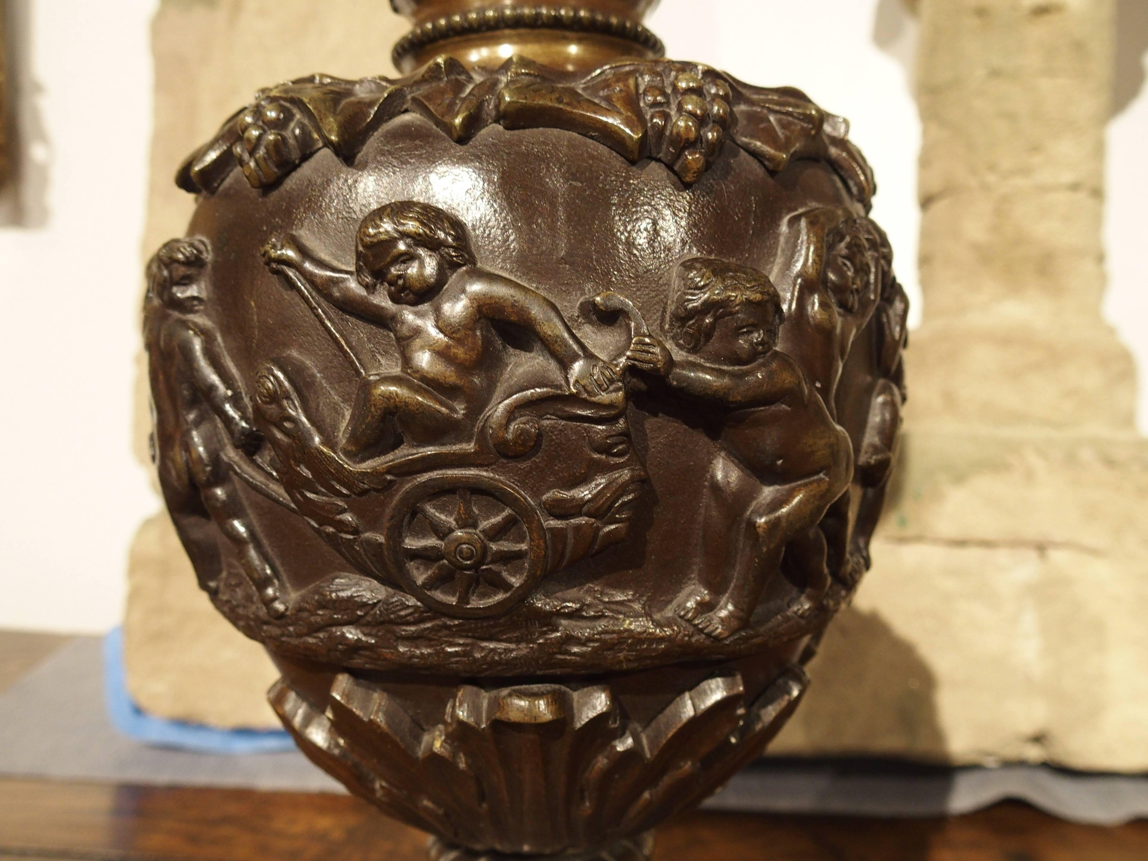 These Renaissance style antique French patinated bronze ewers depict very busy and mischievous Bacchanalian putti at play. A cherub sits atop the handle holding an item in his hands. The musicians are putti with horns, drums and possibly a flute and