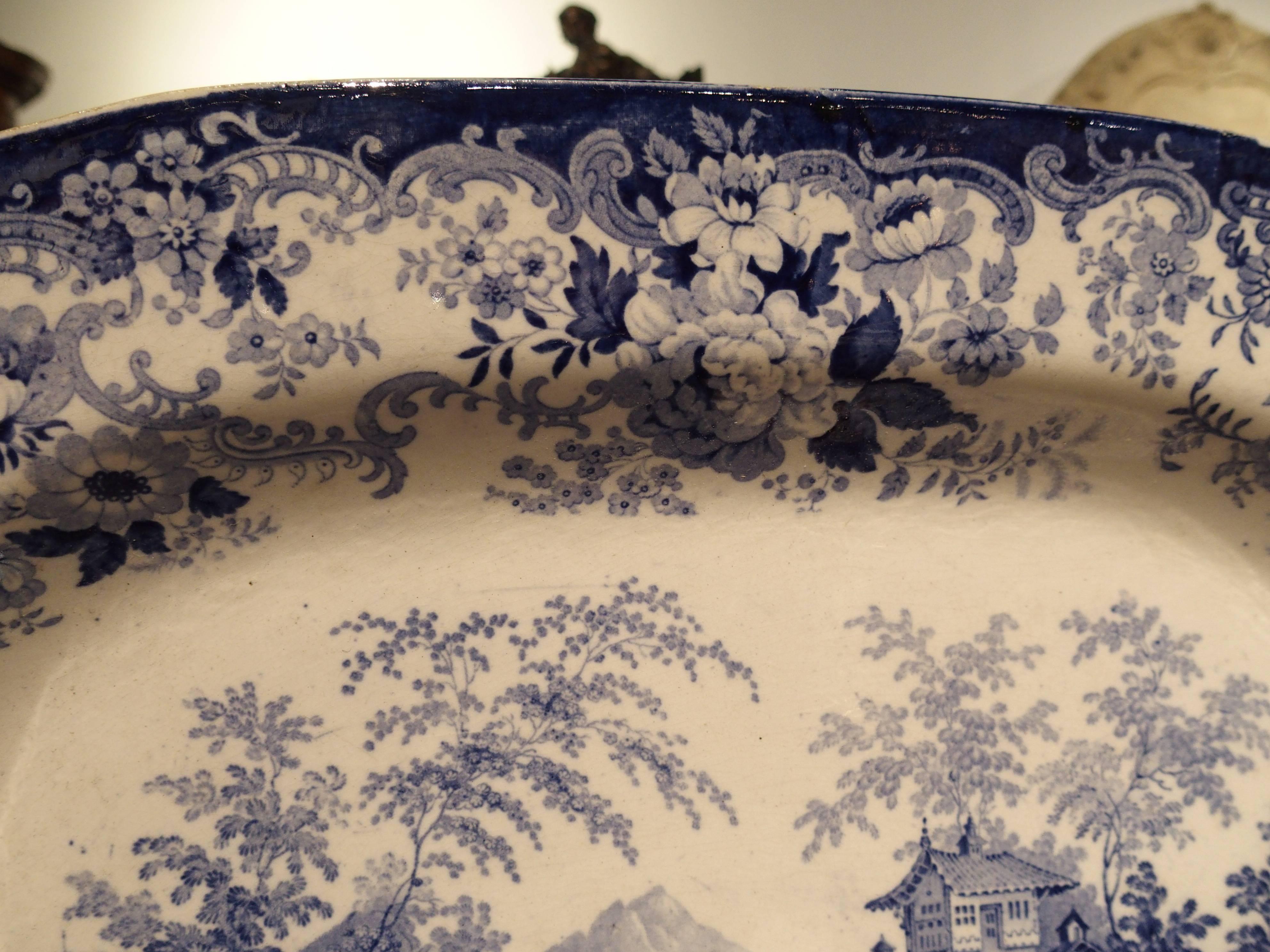 This English blue and white transfer ware platter after Minton’s Genevese pattern, has a charming scene of a lake which is surrounded by homes, mountains and trees, in the chinoiserie style. The border of the platter is a mixture of scrolling floral