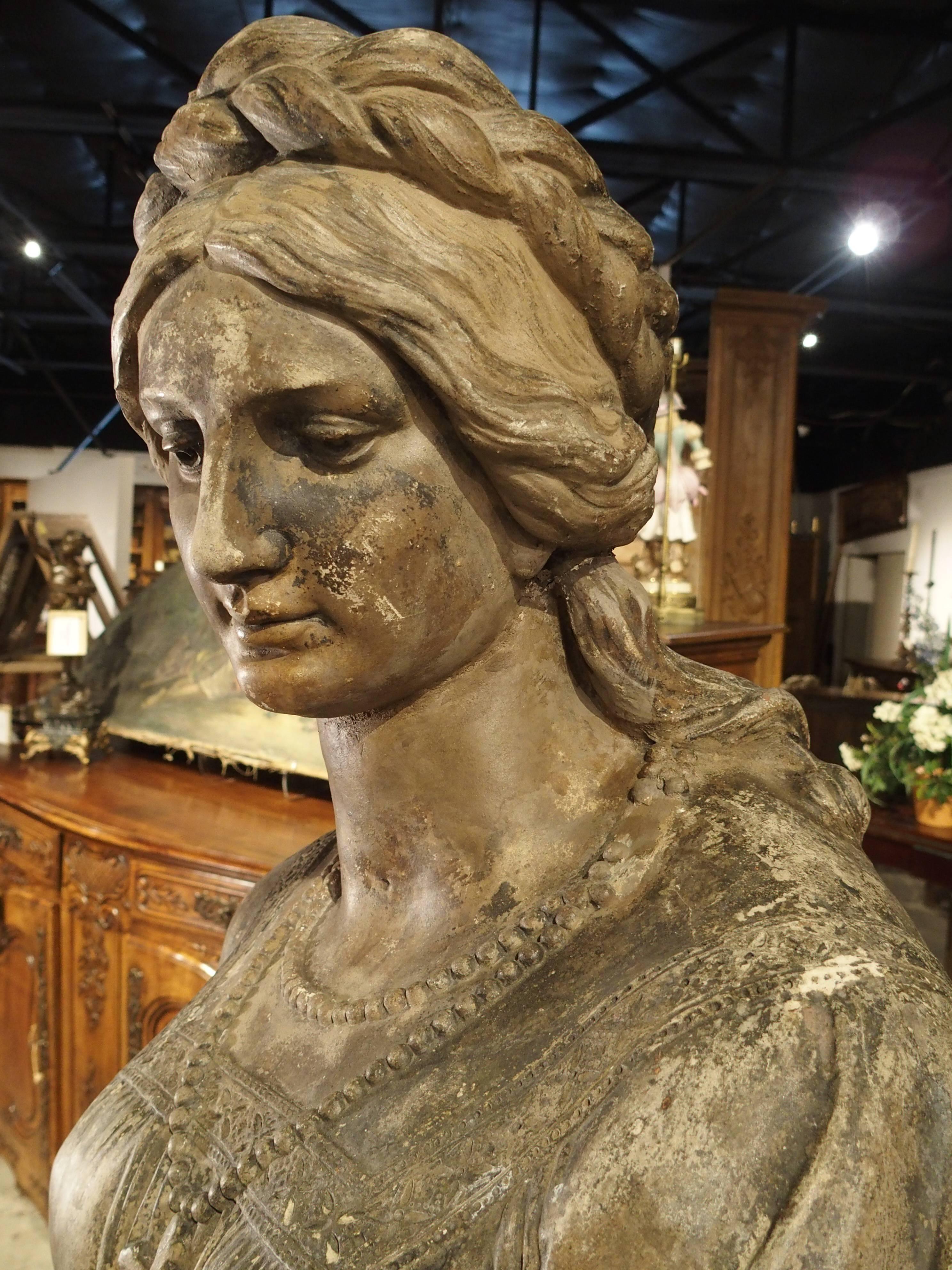 This antique, parcel paint, terra cotta statue depicts a woman carrying a Greek vase with base relief figures on it. She has the front of her long dress draped into her left hand as she steps down from a foliate ledge. Her dress is decorated with