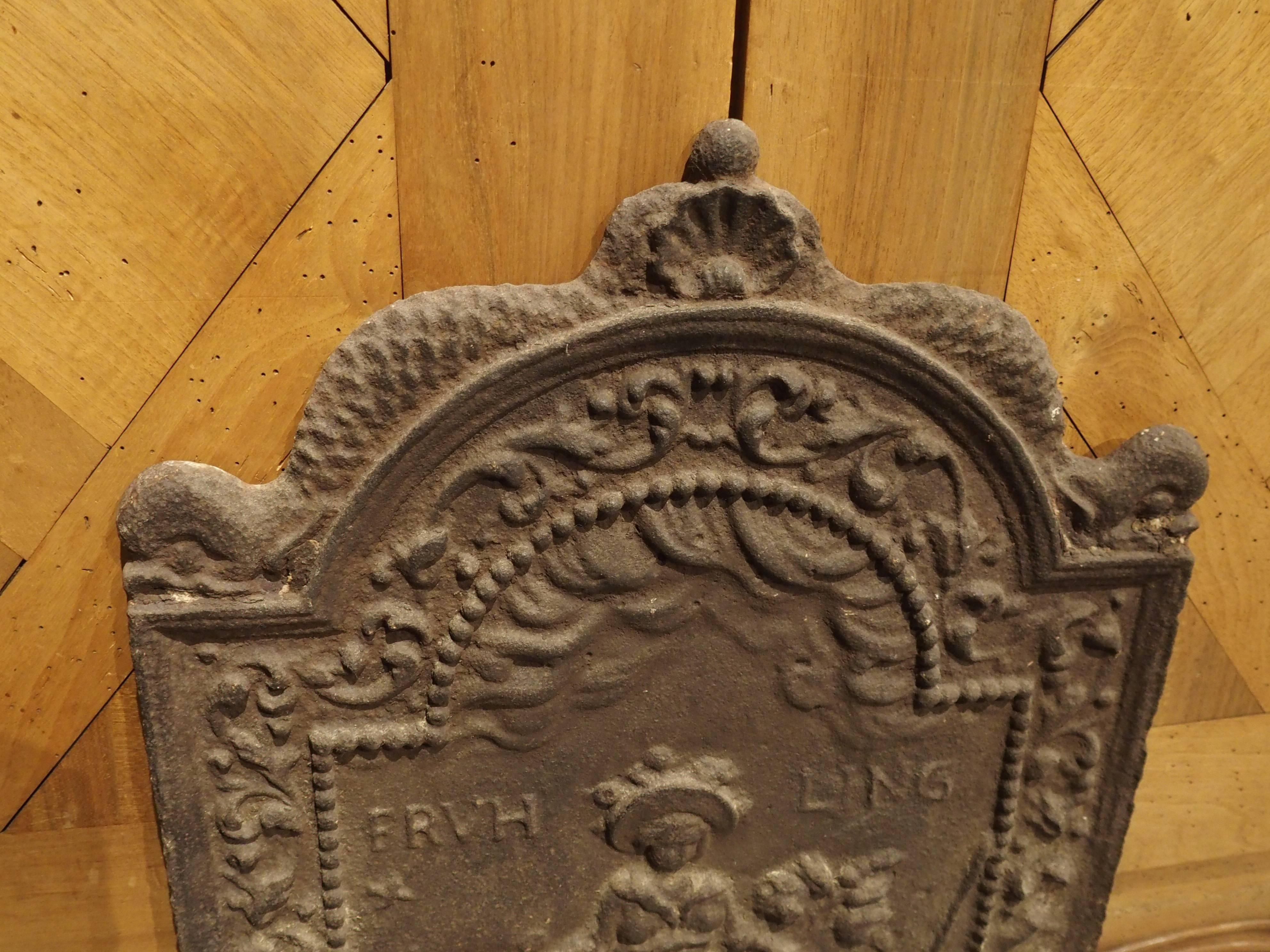 This beautiful 19th century French iron fireback has an arched top with four right angles. At the peak of the arch is a shell surrounded on either side by dolphins. The front depicts a young girl wearing a bonnet in a landscape setting, with a