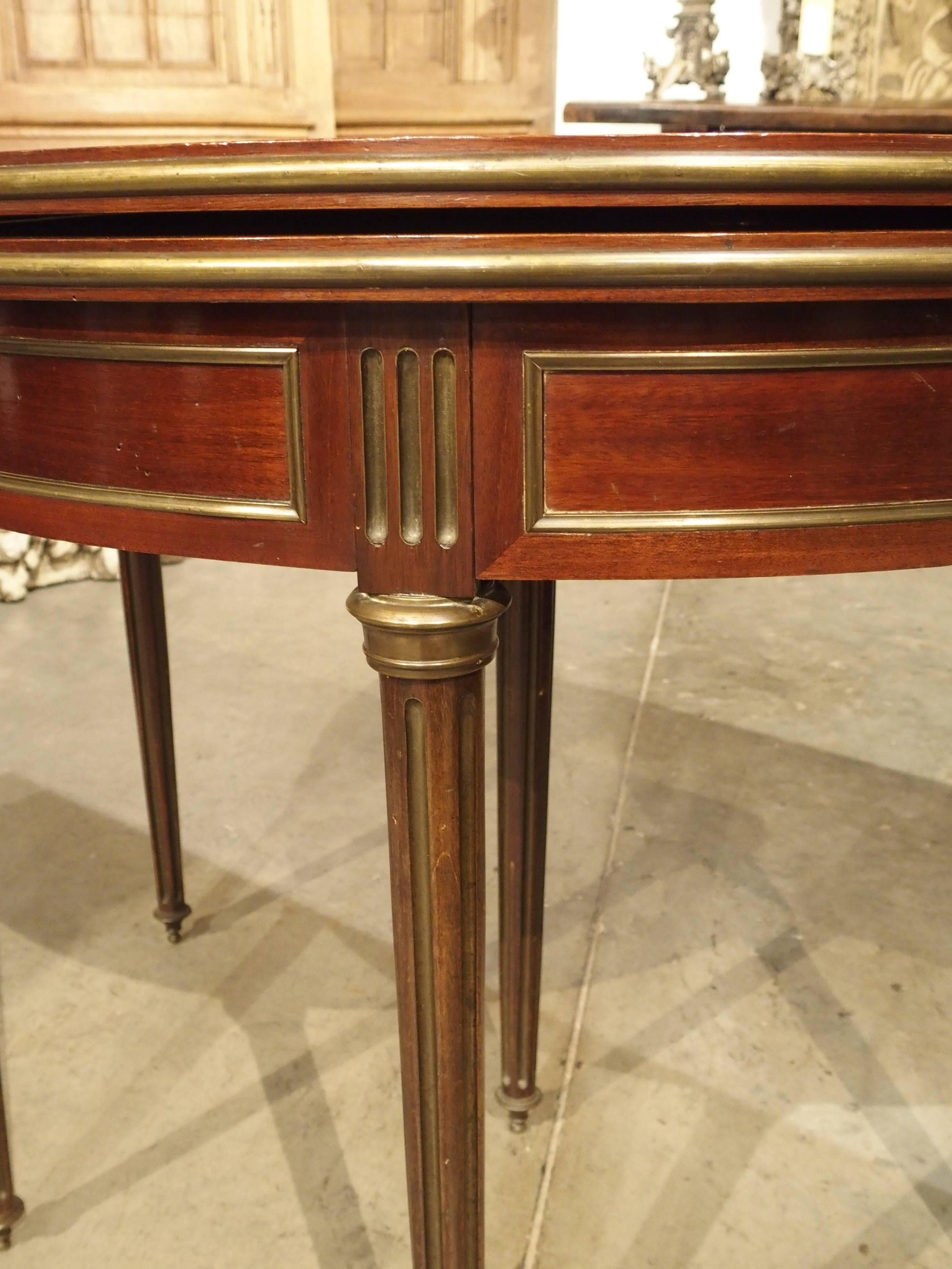 This versatile mahogany and bronze antique French game table can be used closed as a console or open for a game table. It is in the Louis XVI Style and has a half round molding of bronze around the top. The demilune opens to a circle and a felt